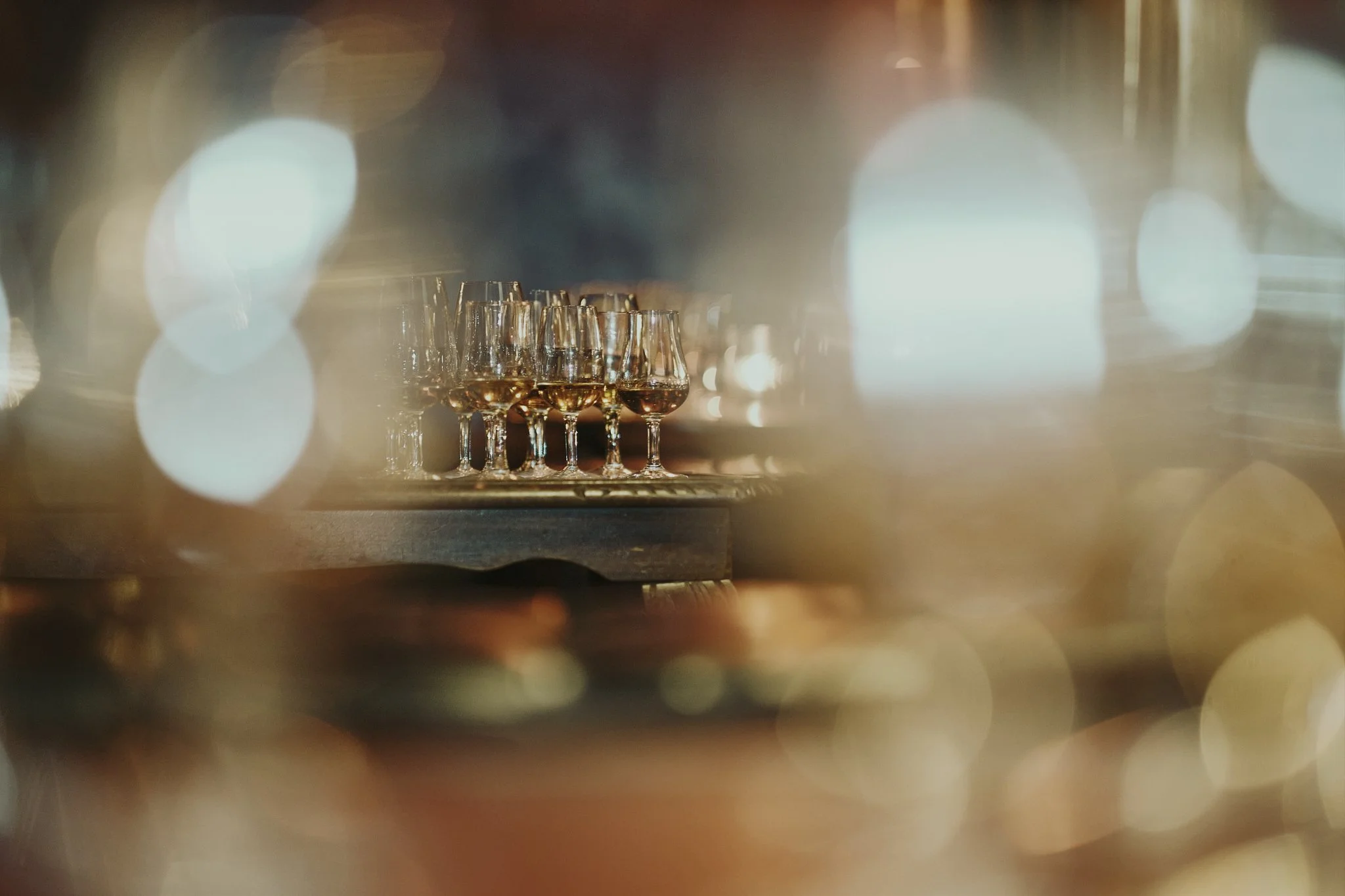 Multiple glasses of white wine on a tray, viewed through a blurred foreground with bokeh lights.