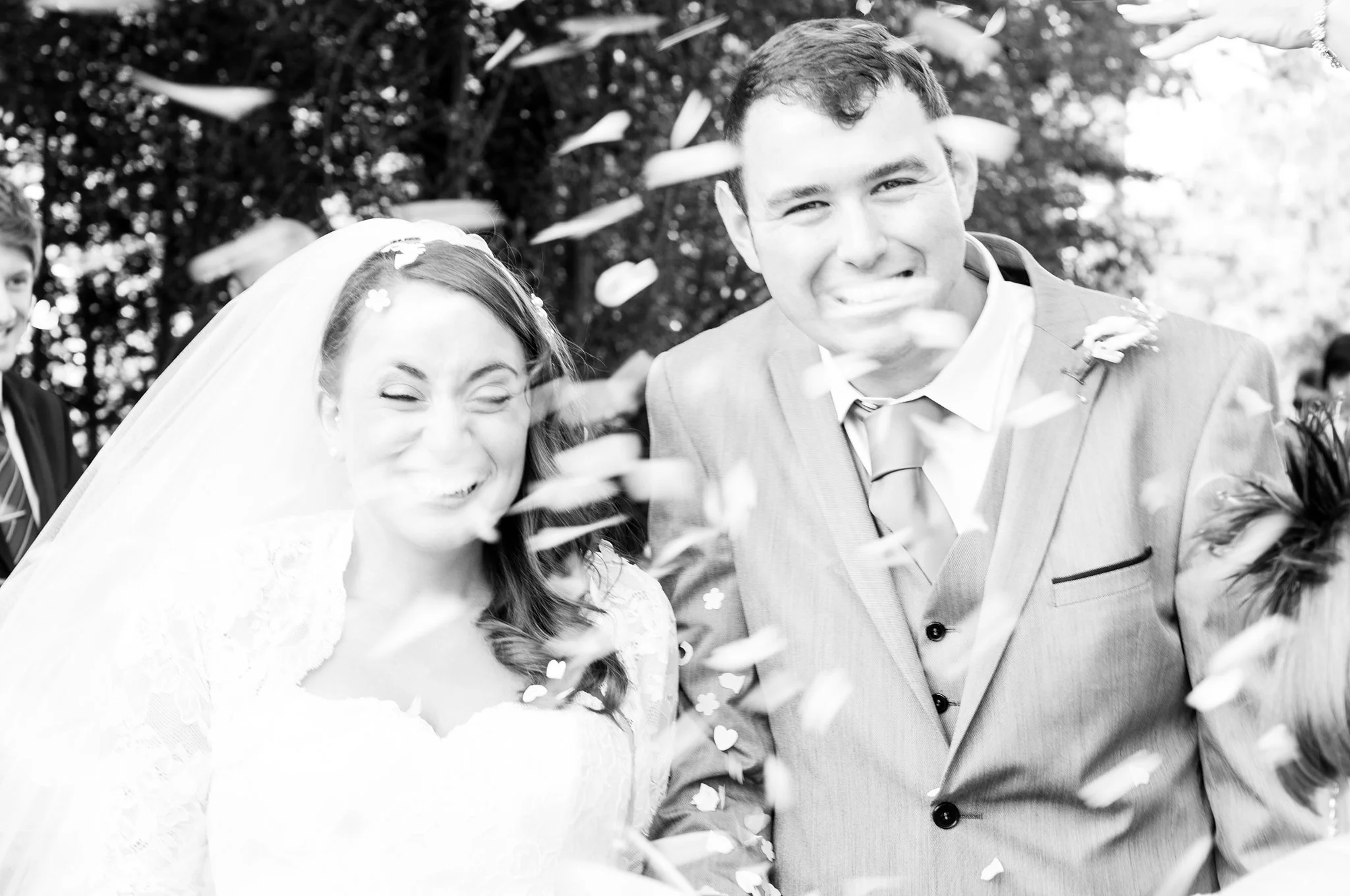 A black-and-white photo of a smiling bride and groom at their wedding, surrounded by falling confetti. The bride is wearing a veil and lace dress, and the groom is in a suit with a tie.