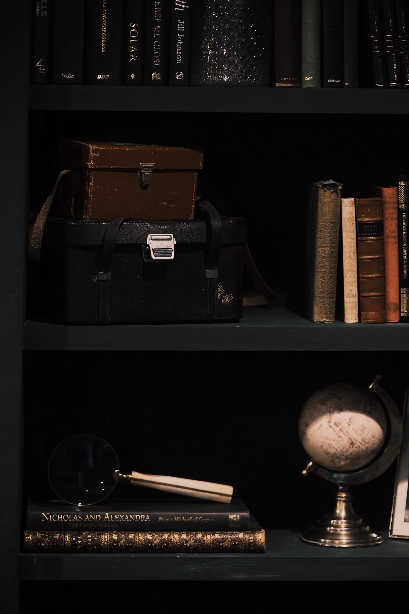 A dark green bookshelf containing books, vintage camera bags, a globe, and a small glass sphere with a white object inside.