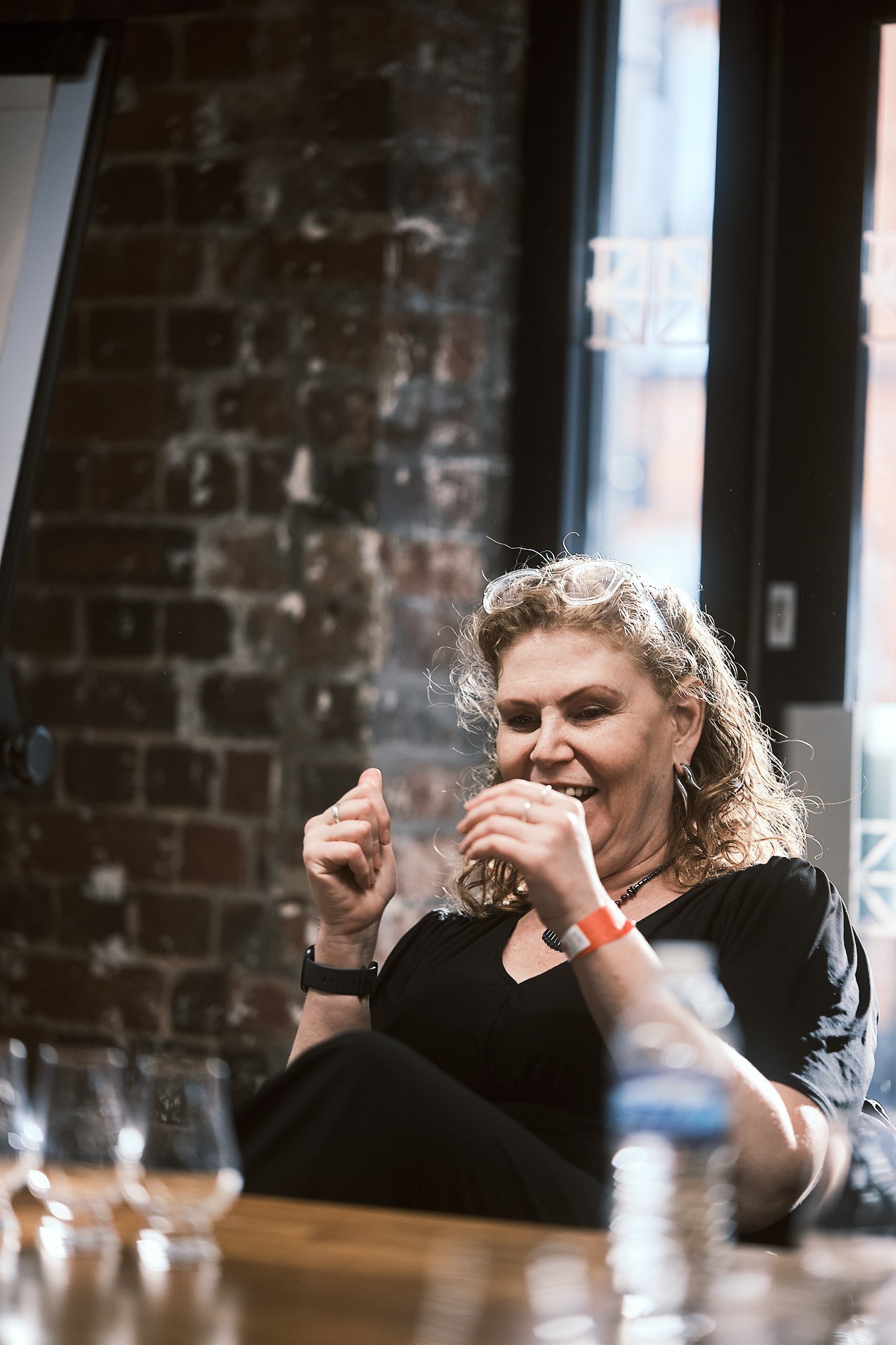 A woman sitting at a table, smiling, with glasses and a watch, in a room with exposed brick wall and large windows.