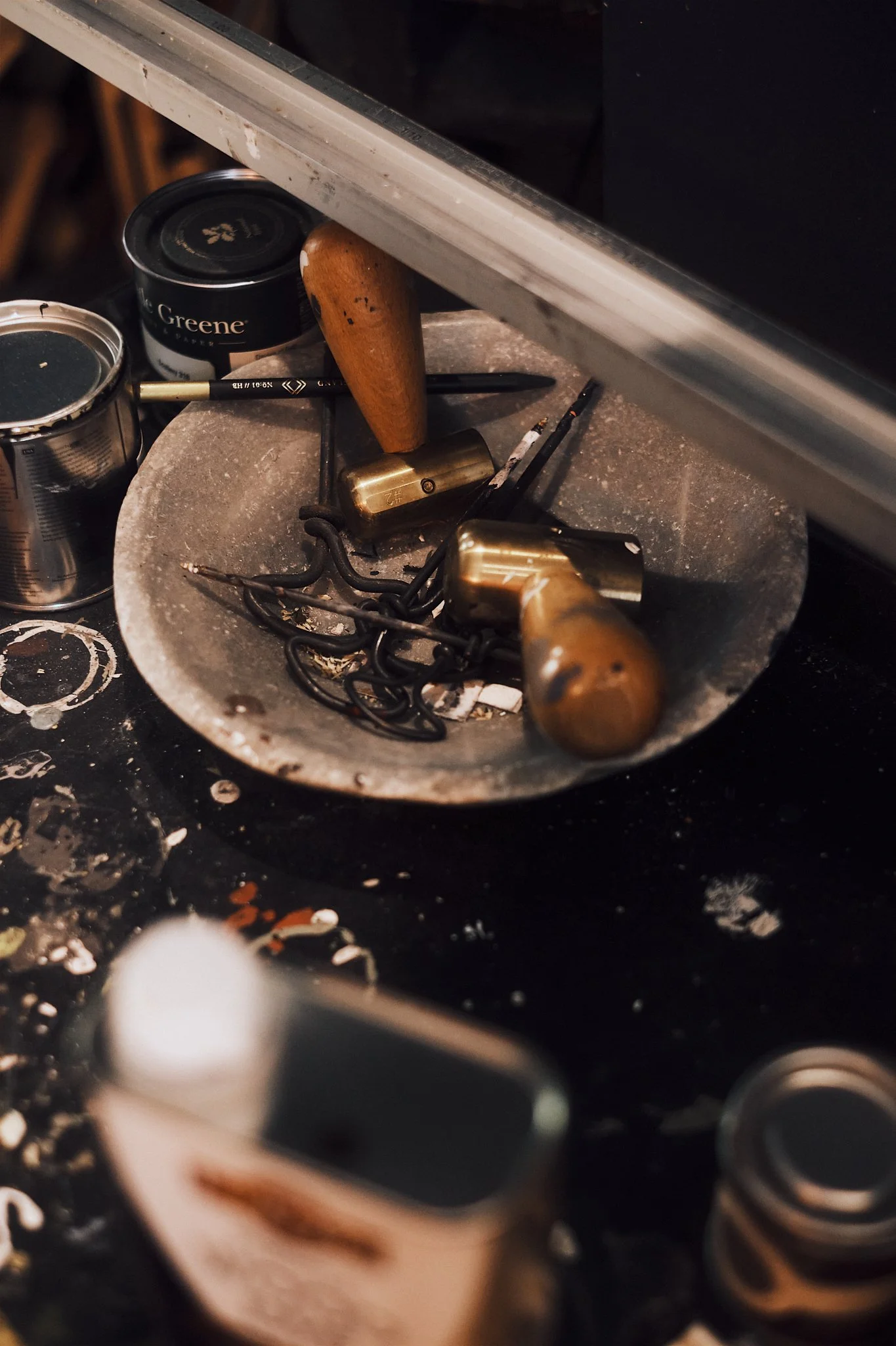A metal work surface with a ceramic or stone bowl filled with small black and brass jewelry tools. Beside it are small tins of paint or finish and a small brush. The work surface has paint splatters and stains.