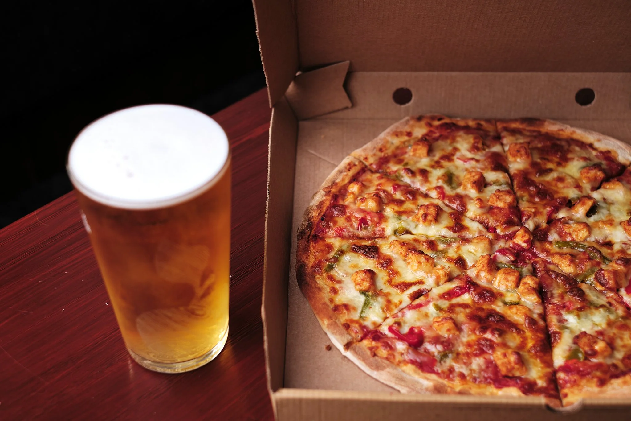 A pizza with cheese, chicken, green peppers, and red onions in an open cardboard box next to a tall glass of beer on a wooden table.