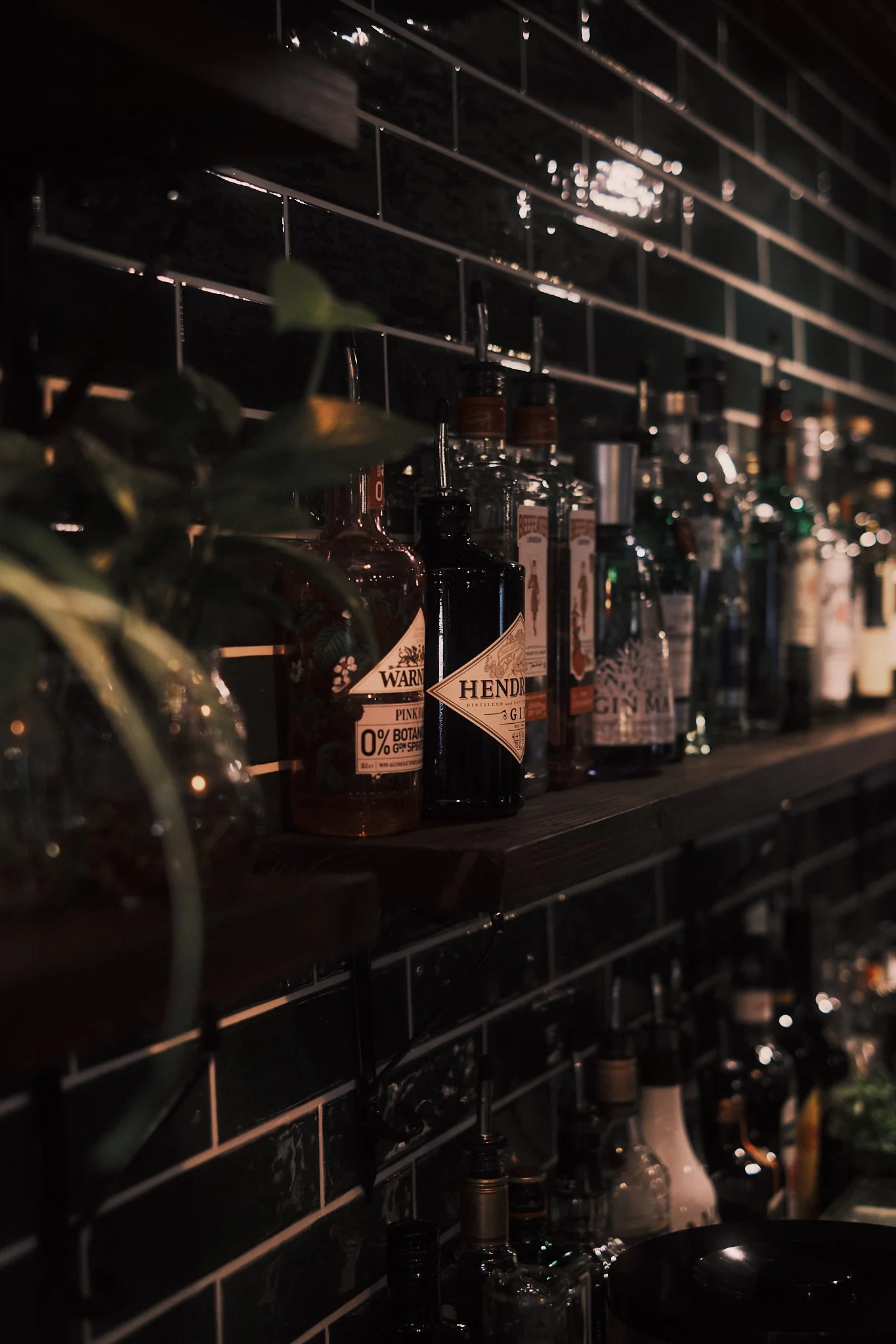 A dark bar shelf with various liquor bottles, including Hendrick's gin, against a black tiled wall.
