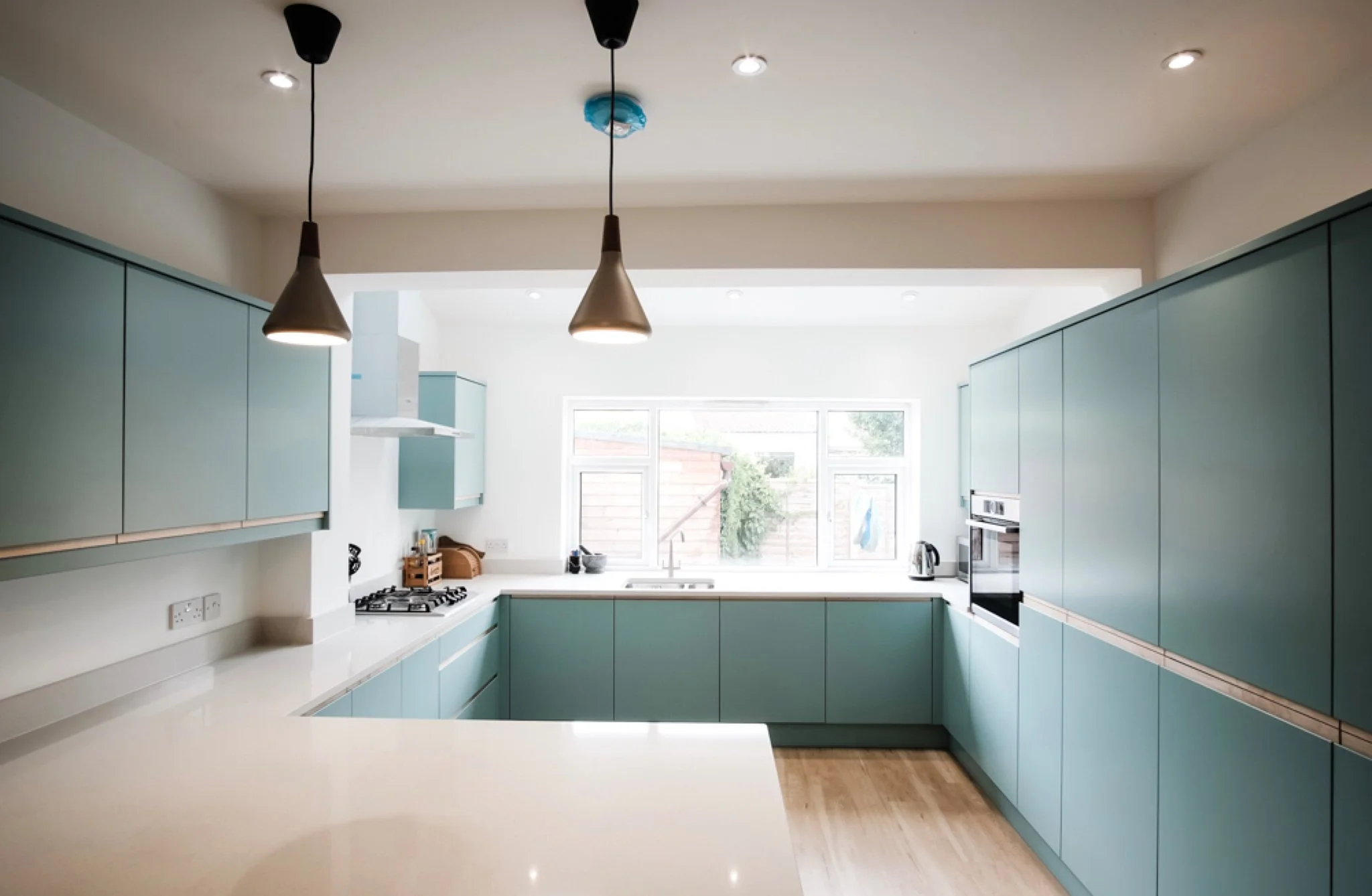 Modern kitchen with light blue cabinets, white countertops, and a large window allowing natural light, with pendant lights hanging from the ceiling.
