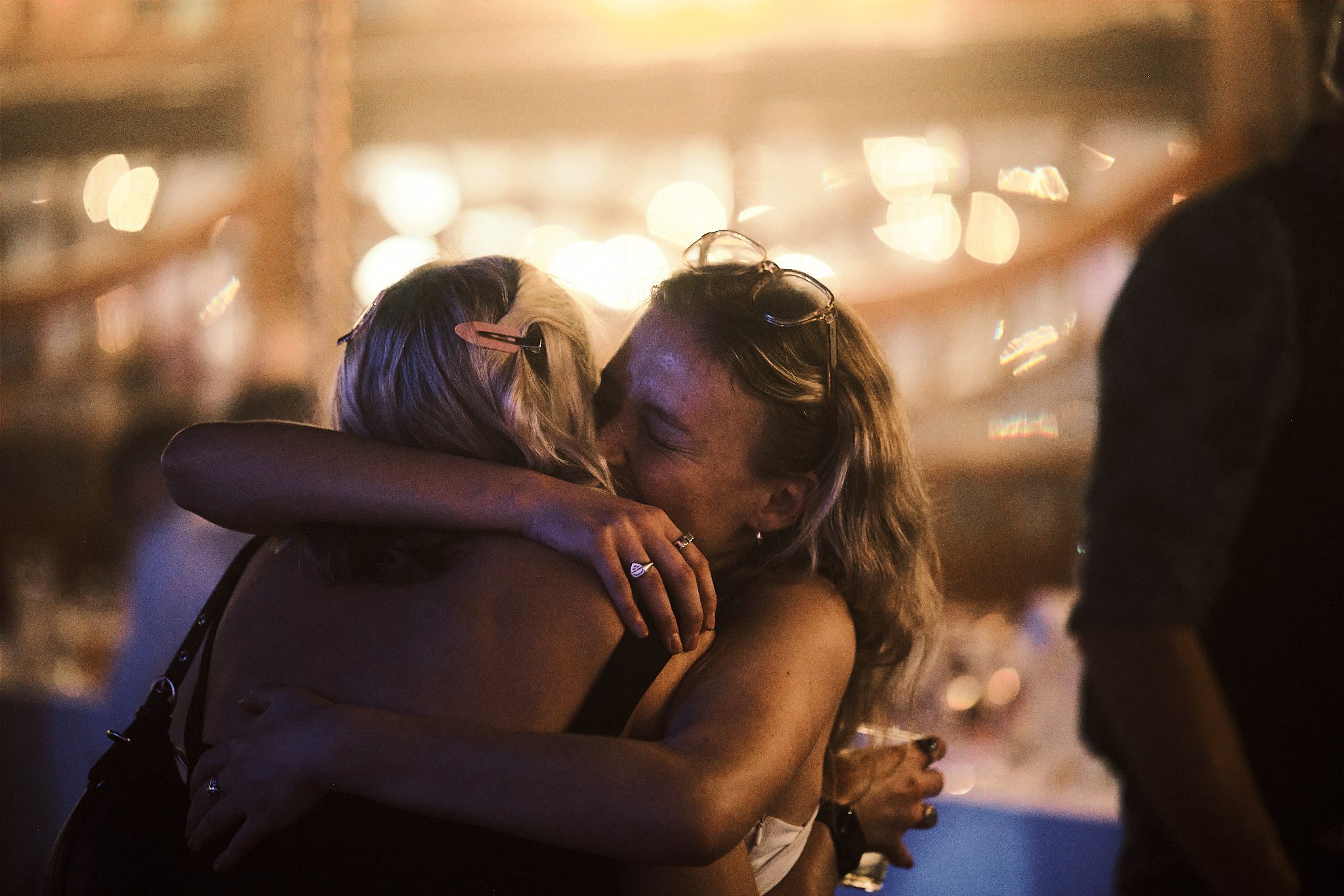 Two women hugging and kissing at a social gathering or party, with blurred lights in the background.