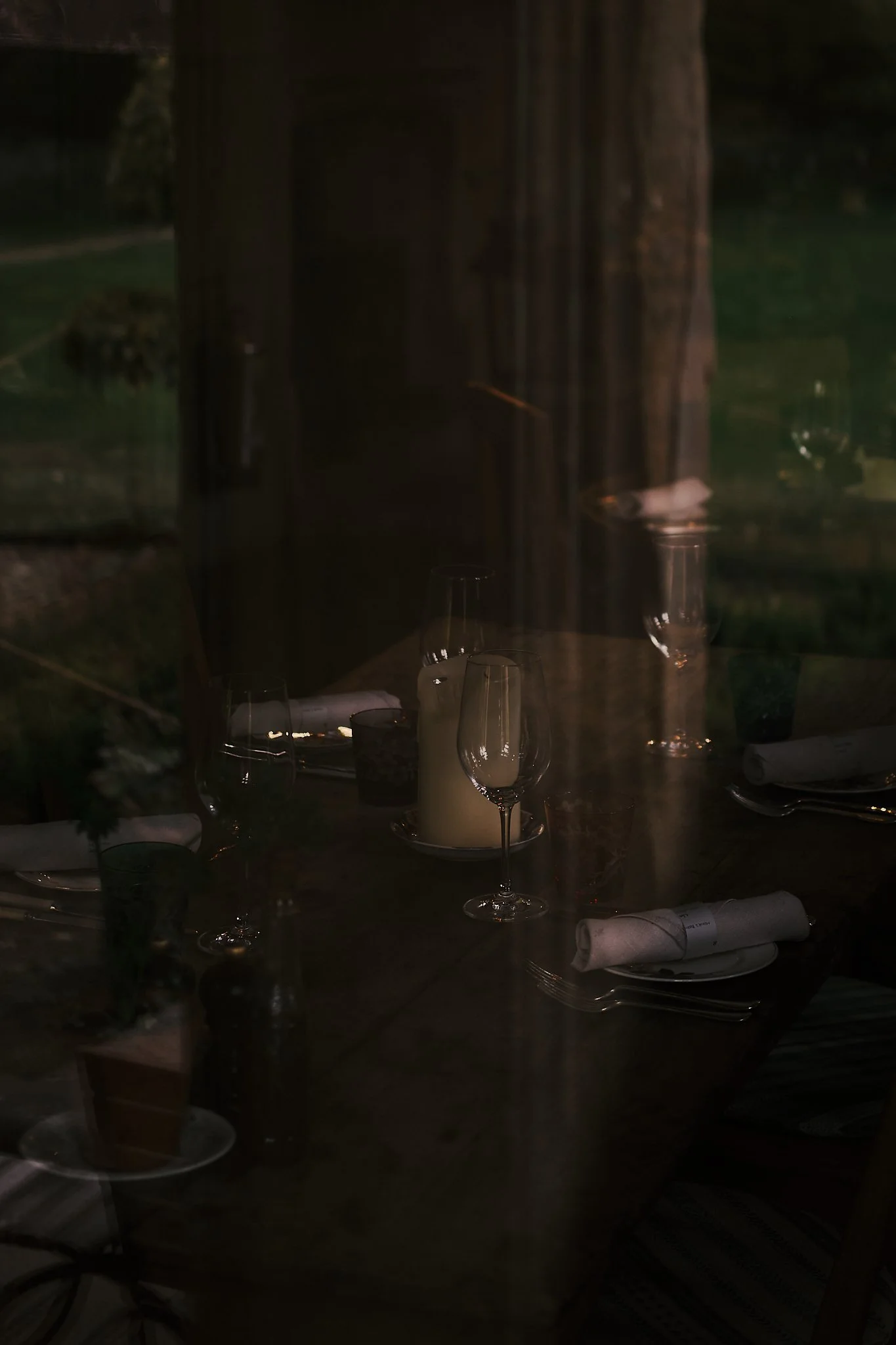 A dimly lit dining table seen through a window, with wine glasses, a candle, napkins, and utensils arranged for a meal. Hotel interior photography at THE PIG-in the Cotswolds