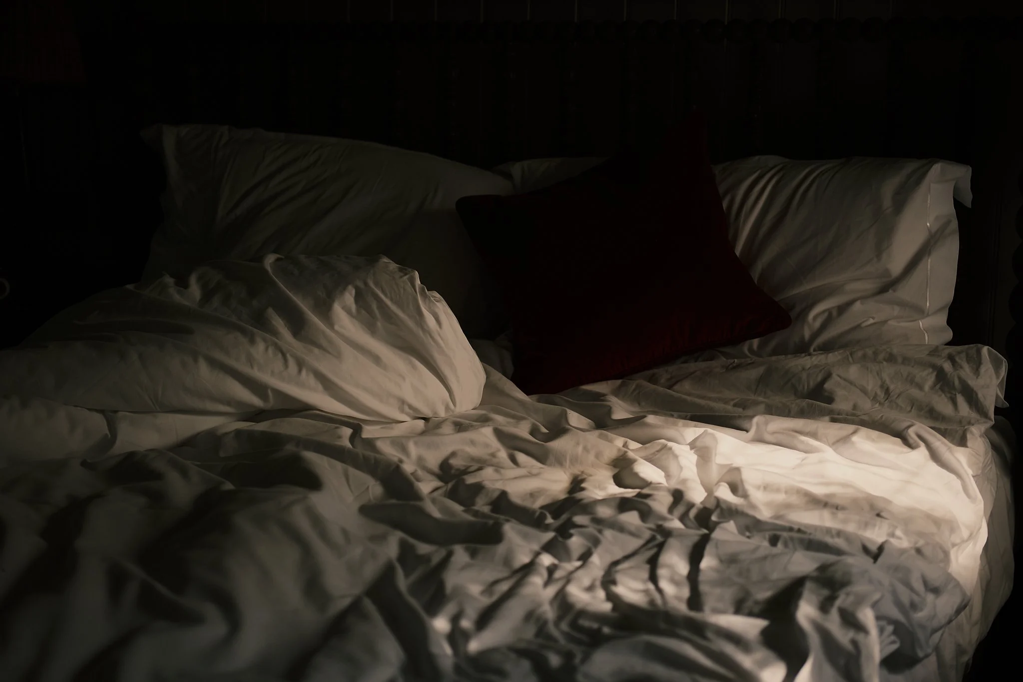 Unmade bed with white sheets and pillowcases and a red pillow, illuminated by a soft light, in a dark room.