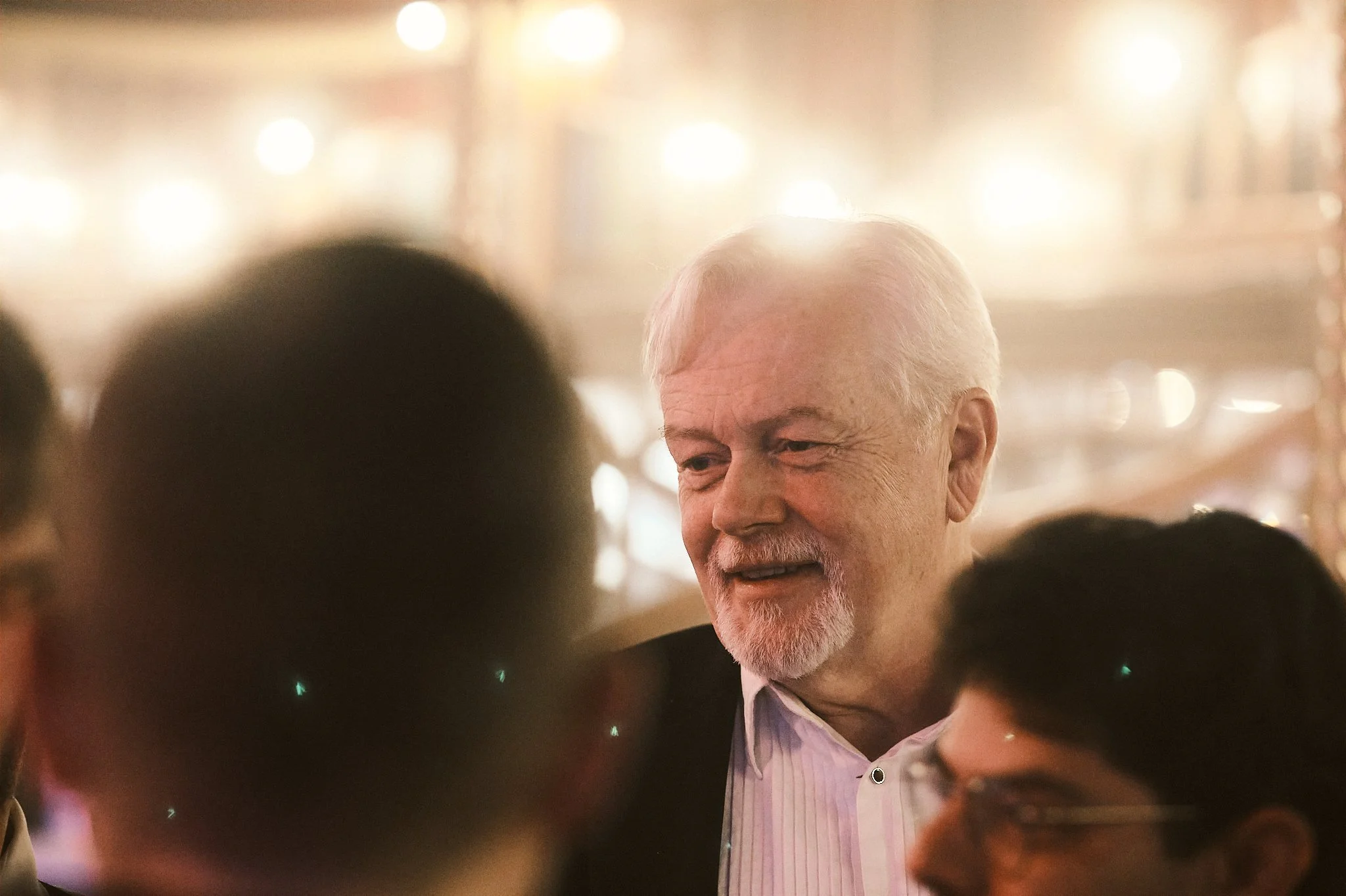 An older man with white hair and a beard, wearing a light pink collared shirt and dark jacket, is talking with a group of people in a warmly lit, lively setting.