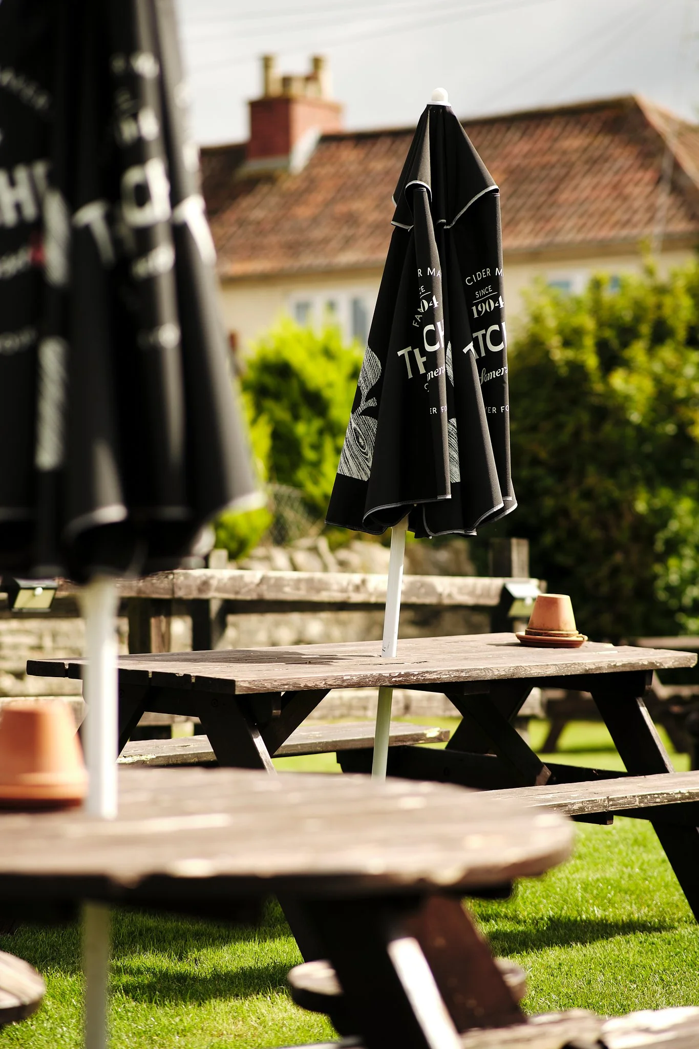Outdoor patio with wooden tables, black umbrellas, one open in the background, and an overturned terracotta pot on one table. Buildings with red roofs and trees are visible in the background.