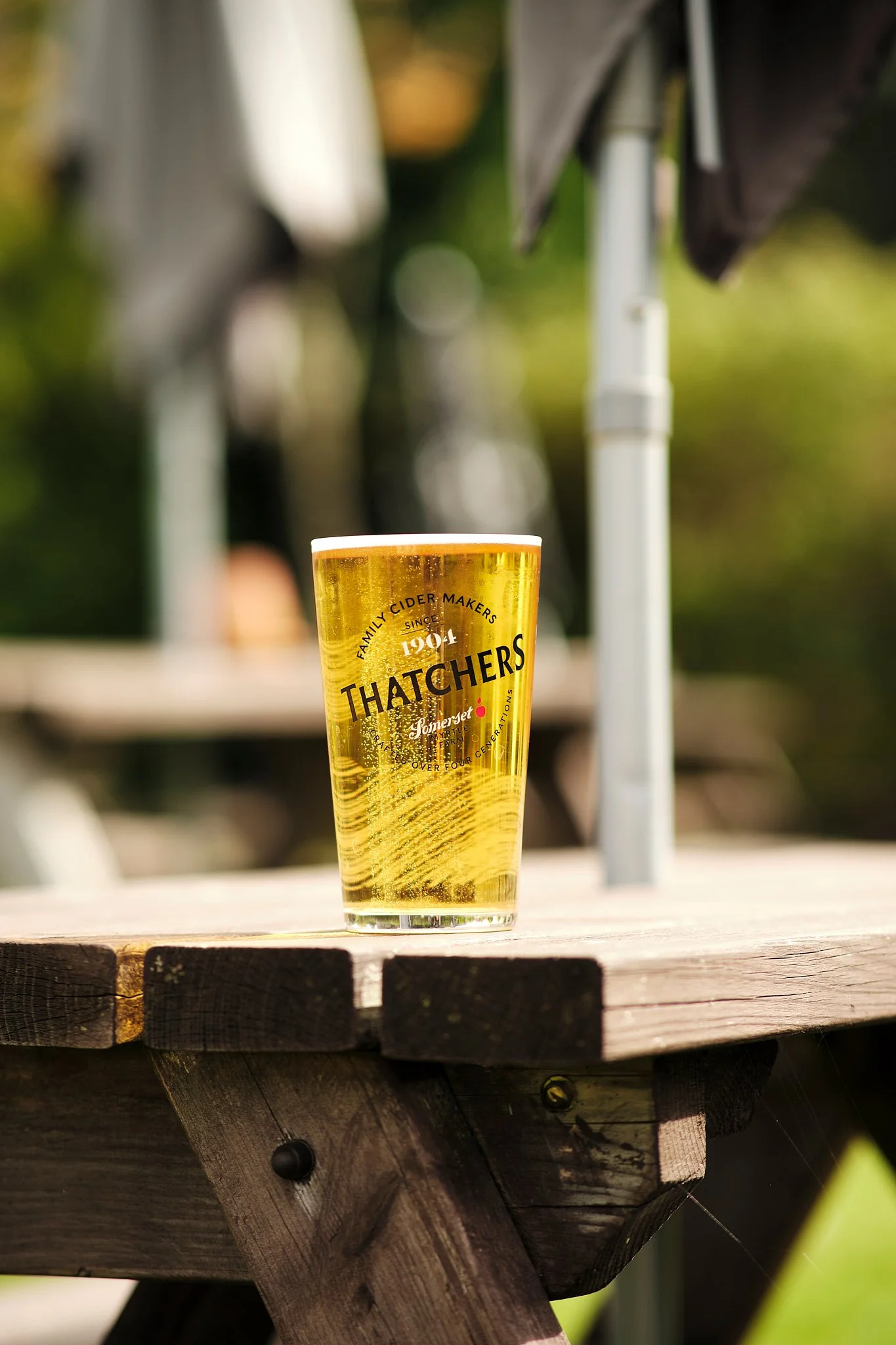 A pint glass filled with golden beer on a wooden table outdoors, with a blurred background and a patio umbrella.