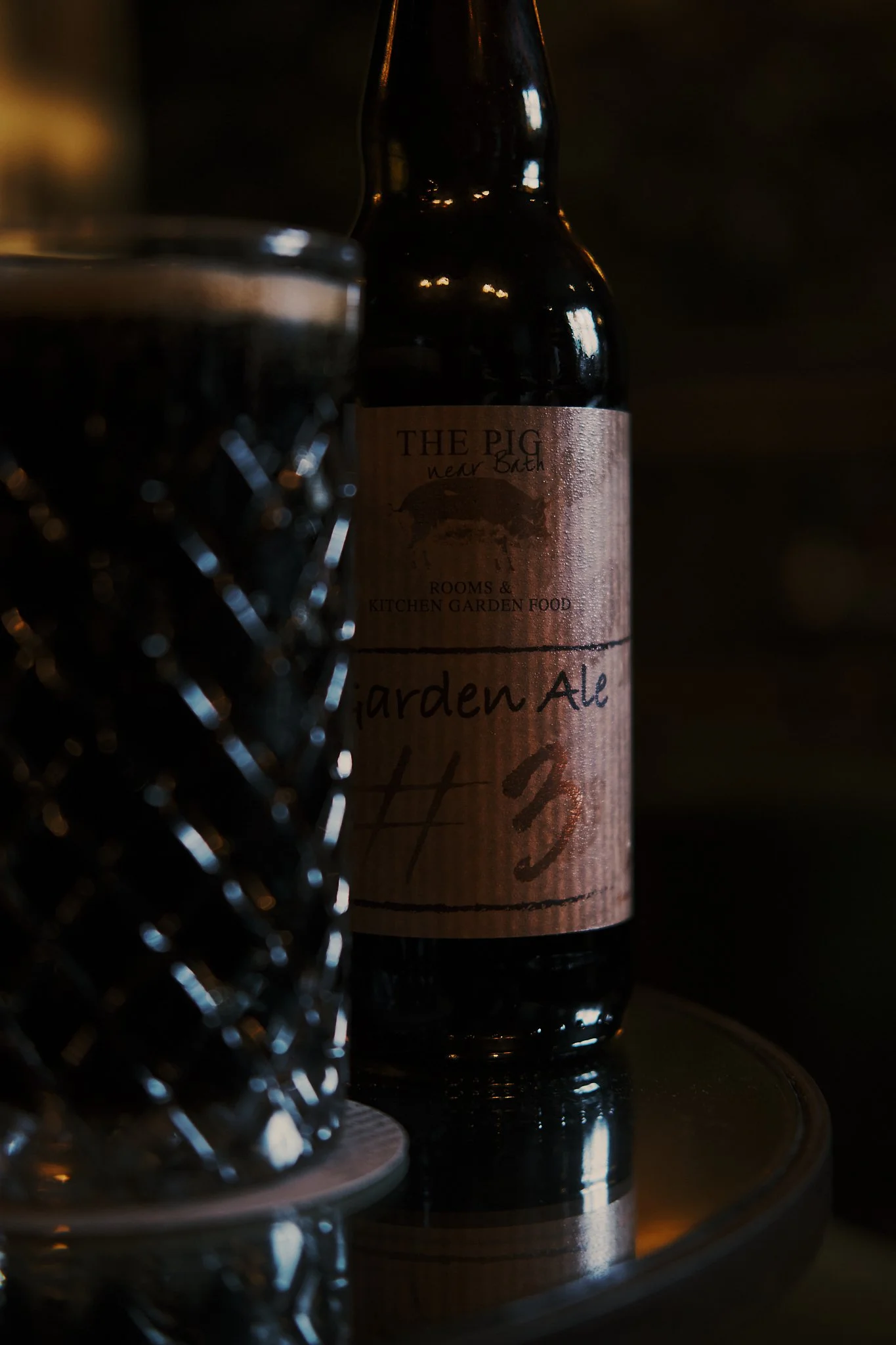 A dark beer bottle labeled 'The Pig' near Bath, with a partially visible glass and reflective surface. Hotel interior photography at THE PIG-in the Cotswolds