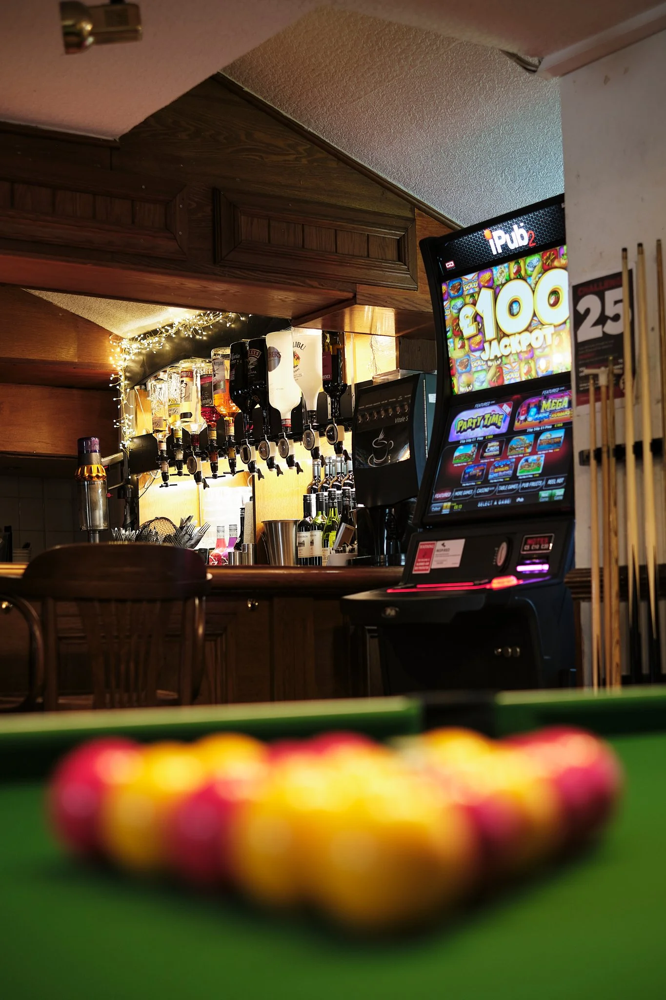A bar area with liquor bottles hanging and stacked behind the counter, next to a lottery and slot machines, with pool balls on a pool table in the foreground.