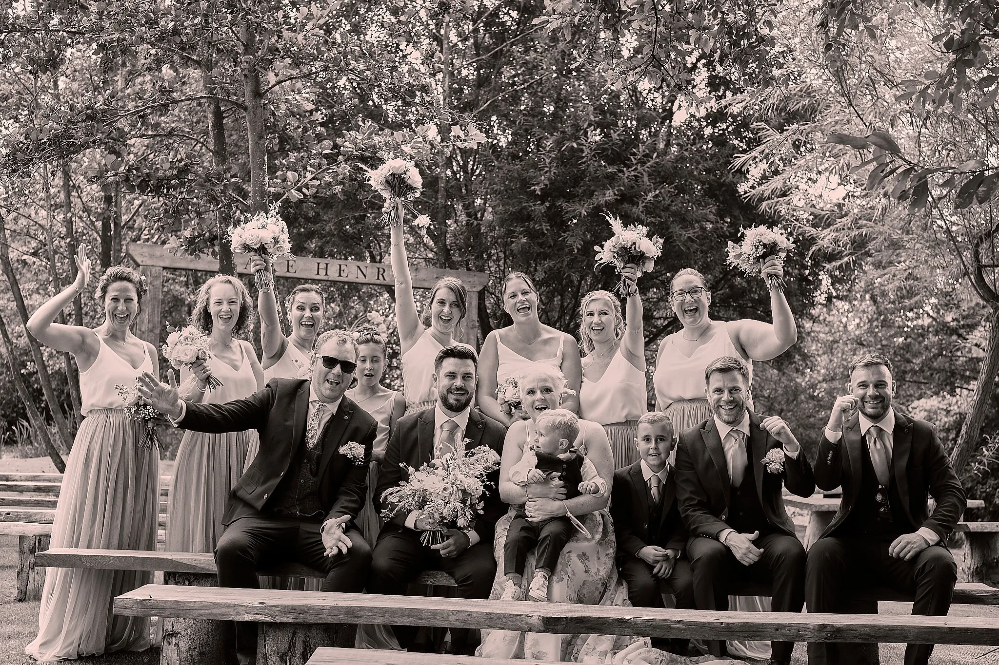 Group of people at a wedding celebration, posing on outdoor steps with trees in the background, some holding bouquets, all smiling and making joyful gestures.