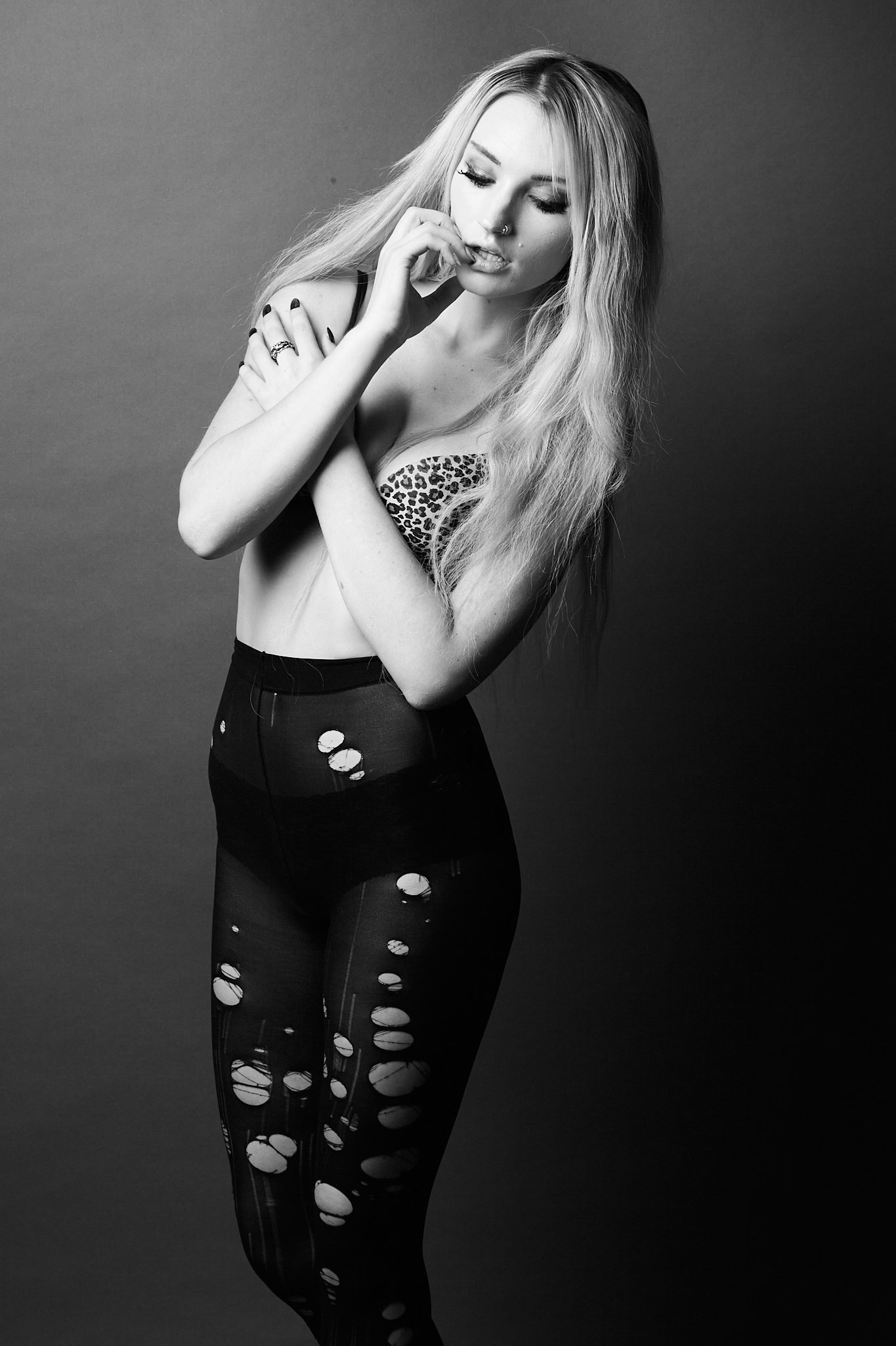 A young woman in black ripped stockings, leopard print top, and long wavy hair, standing against a dark background, with one hand near her face and the other crossed over her chest.