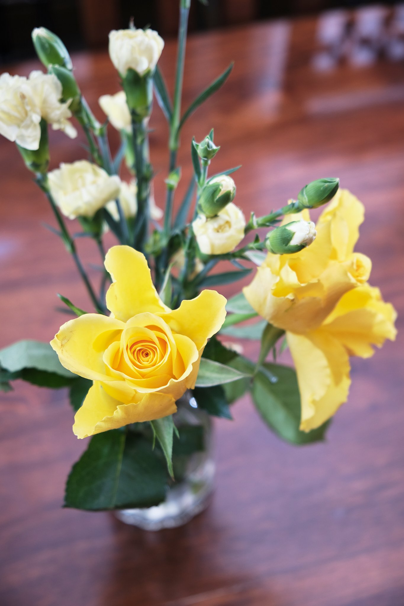 Close-up of a yellow rose and white flowers in a small glass vase on a wooden surface.