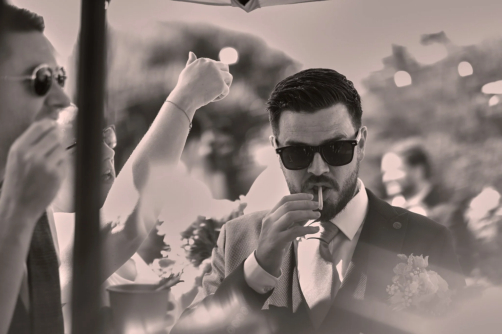 A black and white photo of a man in sunglasses, wearing formal attire, brushing a woman's hair or head, with a blurred background of trees. The woman’s arm is raised, and she appears to be holding a cup. The scene is outdoors with soft lighting.
