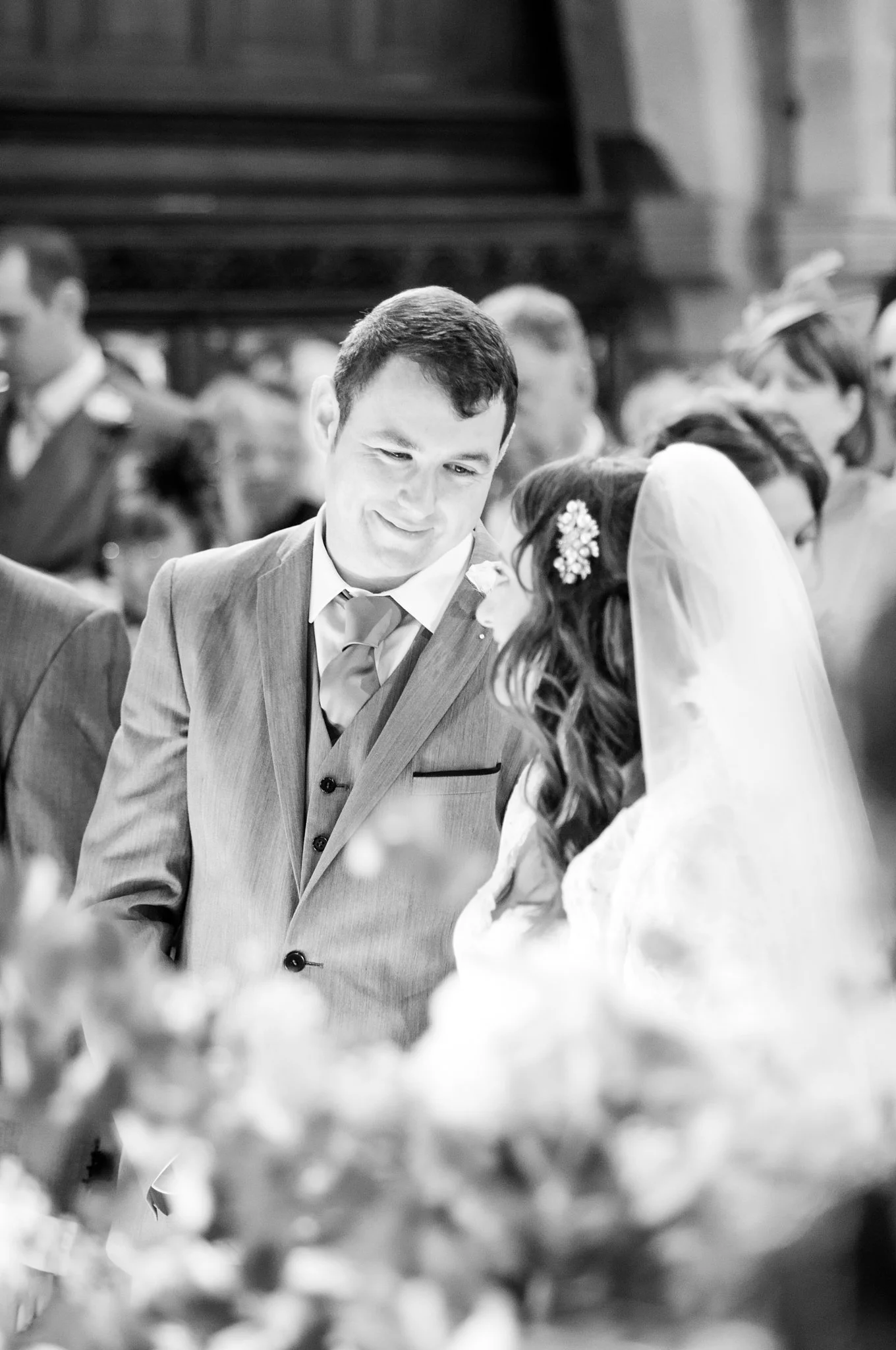 Black and white photo of a groom and bride during a wedding ceremony, focusing on the groom smiling and looking at the bride, with guests in the background.
