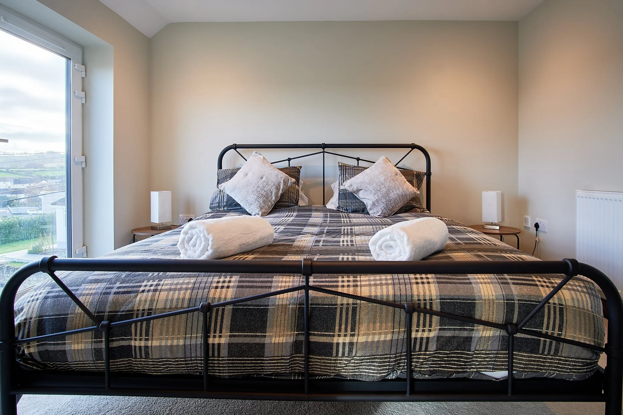 A neatly made bed with a black metal frame, two rolled white towels on top, and four pillows with striped and velvet covers, in a room with beige walls, a side table with a lamp on each side, and a large window with a view of a cloudy sky and houses 
