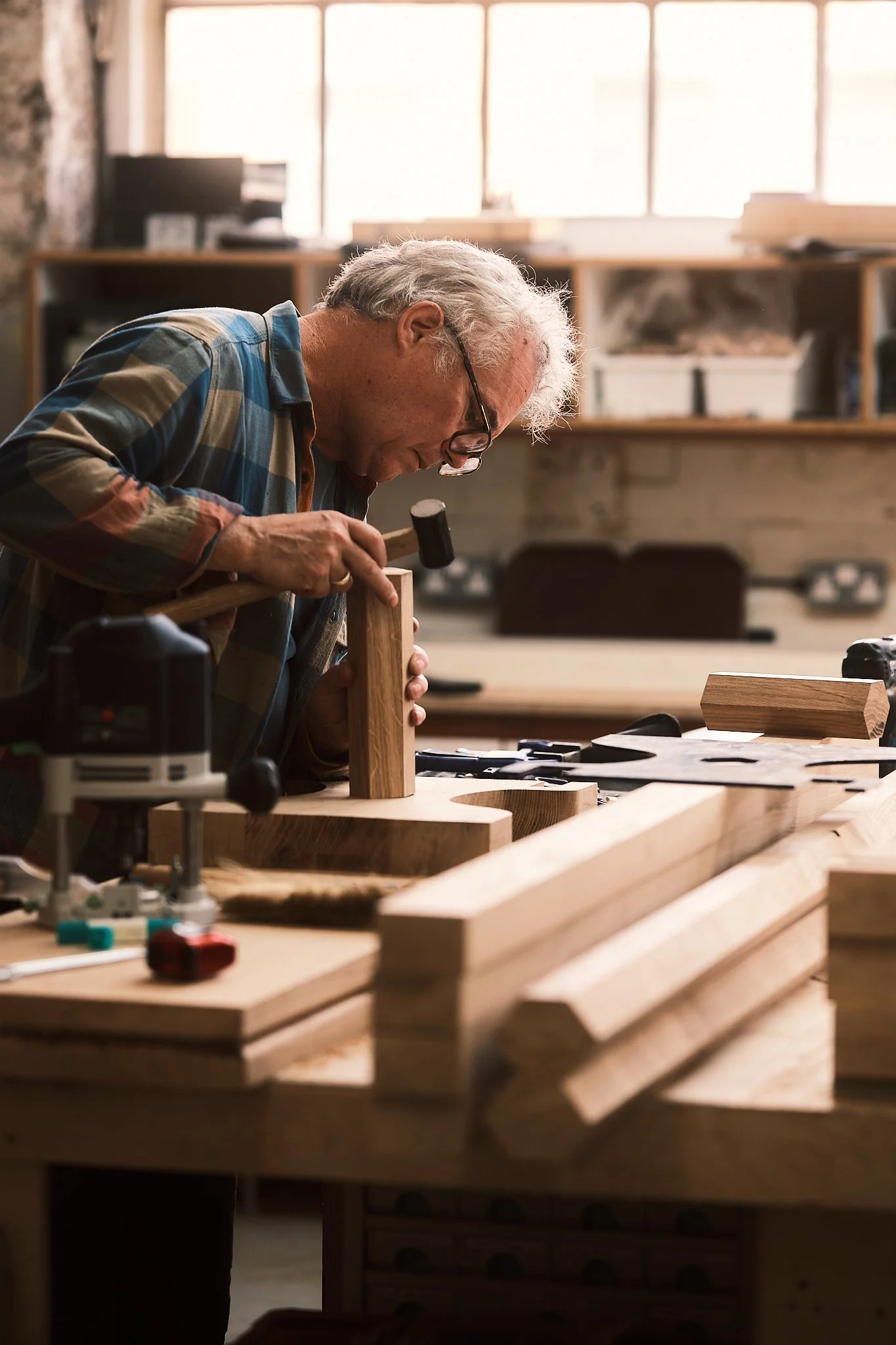 An elderly man with gray hair, glasses, and a plaid shirt is working in a wood workshop, hammering a piece of wood on the worktable.