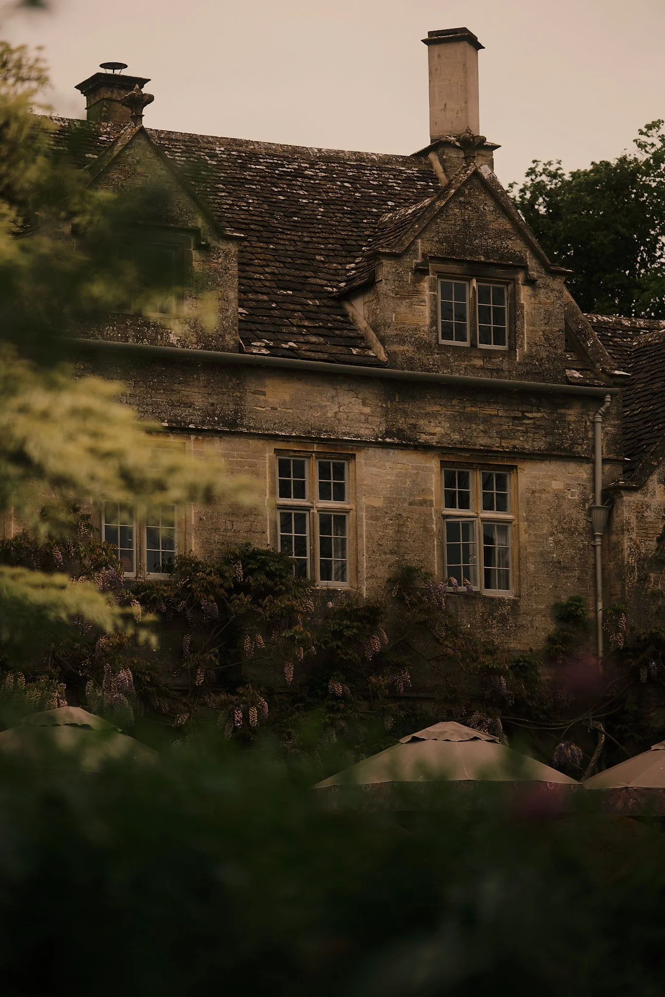An old stone house with multiple chimneys and windows, partly obscured by foliage and purple flowers, under a cloudy sky during dusk. Hotel interior photography at THE PIG-in the Cotswolds