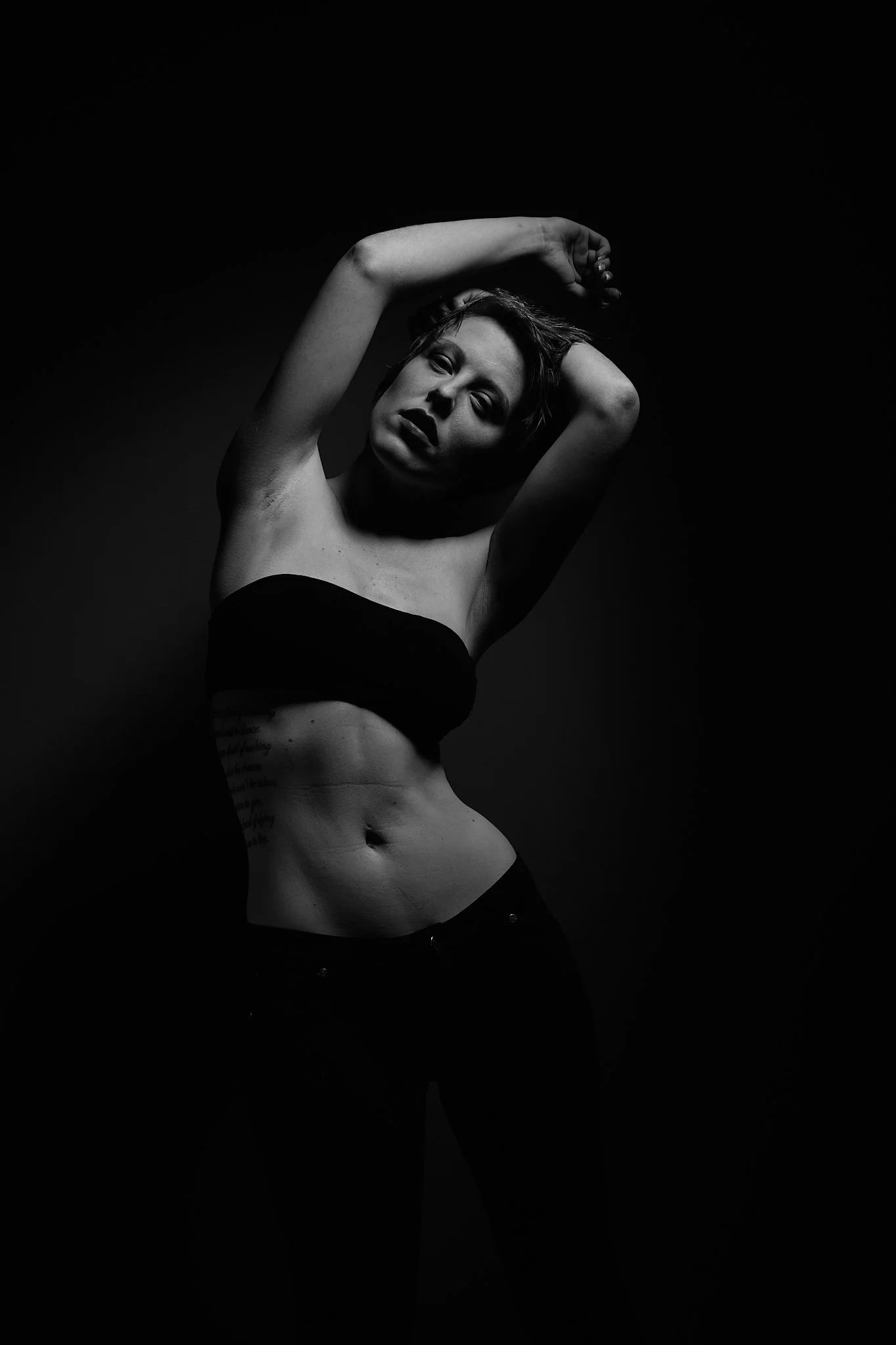 A woman with short hair posing with her arms above her head in a black tube top and pants, in a high-contrast black-and-white photograph.