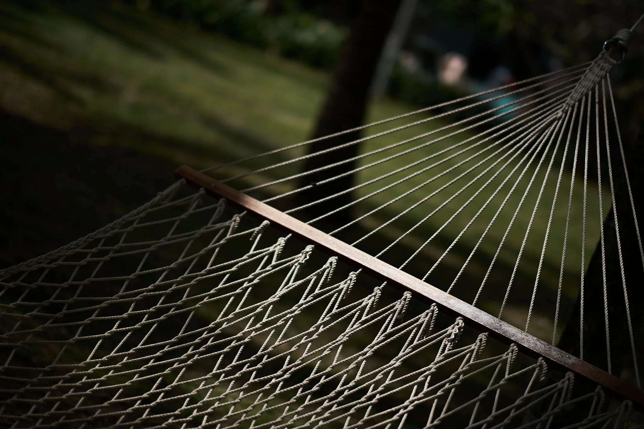 Close-up of a hammock made of ropes with a wooden support, set outdoors with sunlight and greenery in the background.