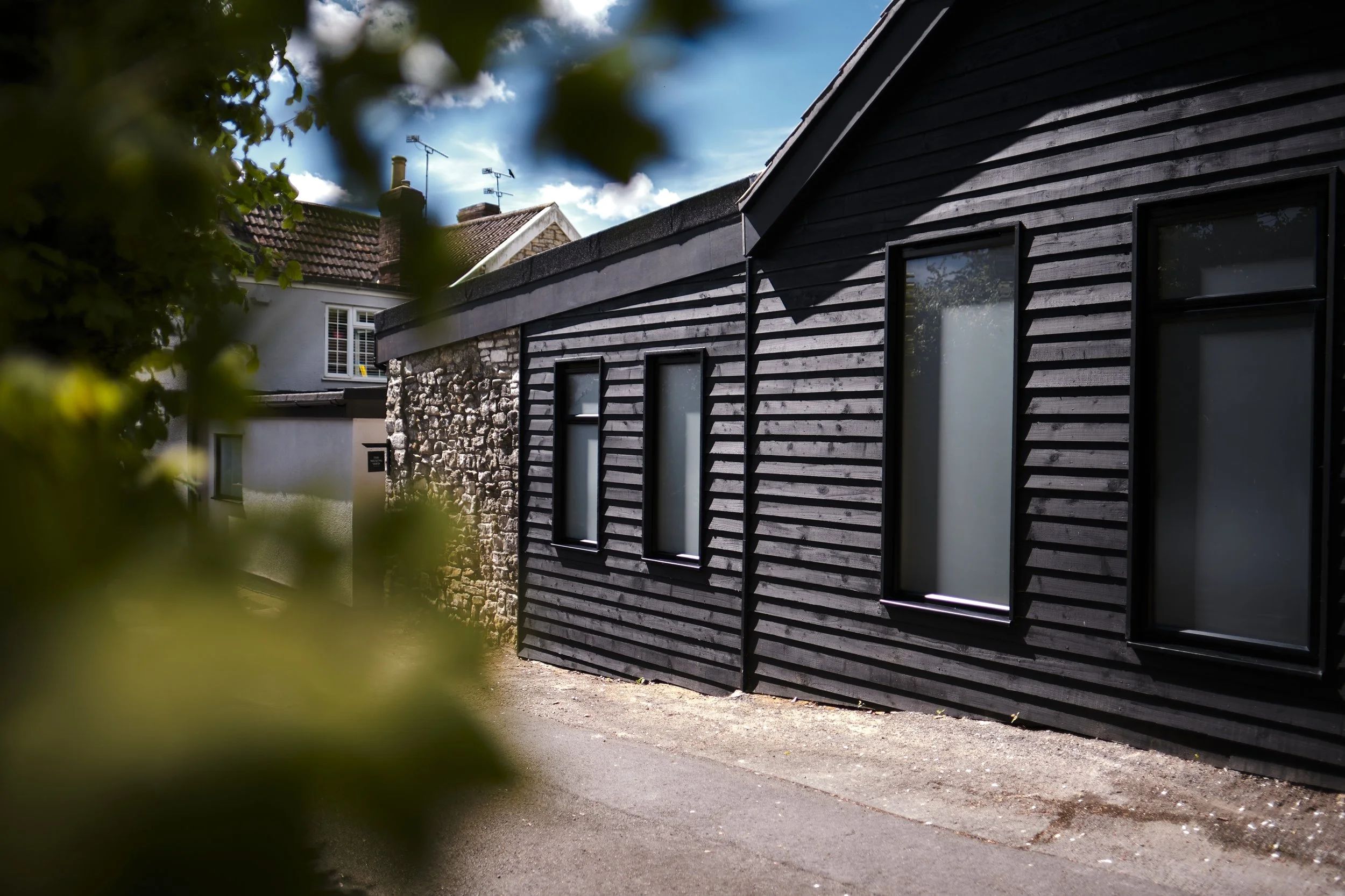 Modern house with black wooden siding and multiple rectangular windows, seen partially through foliage, with neighboring house and clear sky in background.