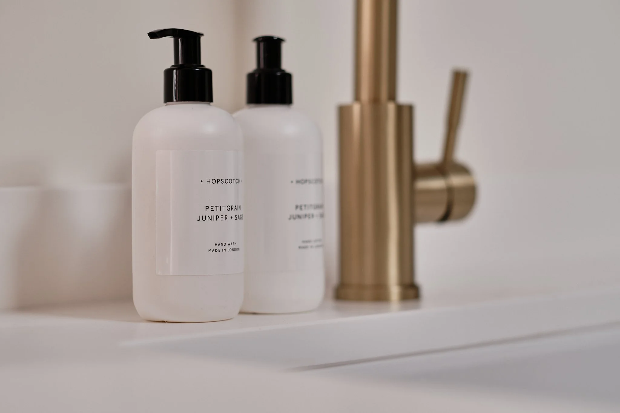 White bottles of Hopscotch hand wash labeled Petitgrain, Juniper, and Sage on a white bathroom counter, with a gold faucet in the background.