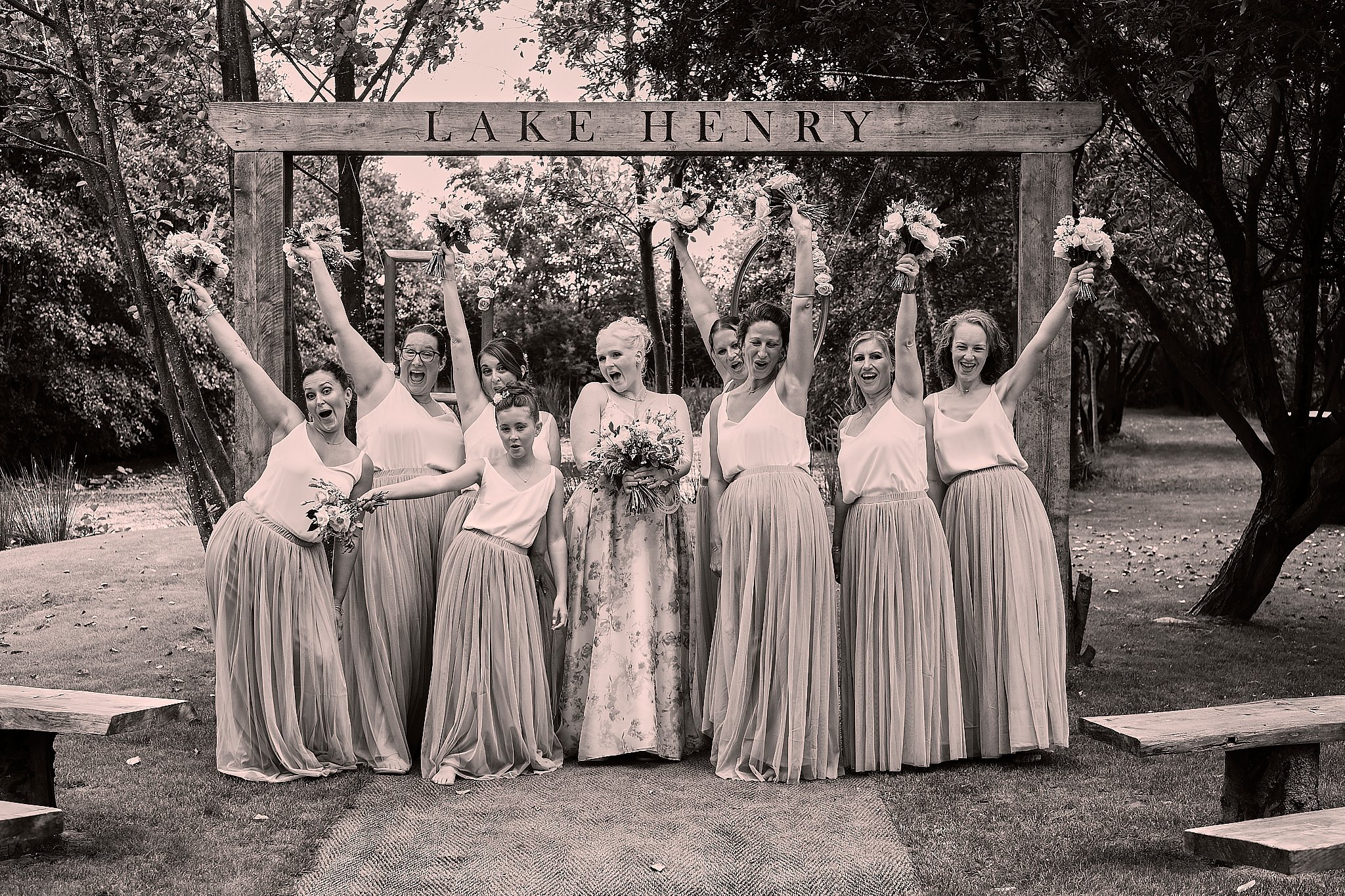 A group of women in bridesmaid dresses standing under a wooden arch with a sign that reads 'Lake Henry'. They are holding bouquets and celebrating at a wedding.
