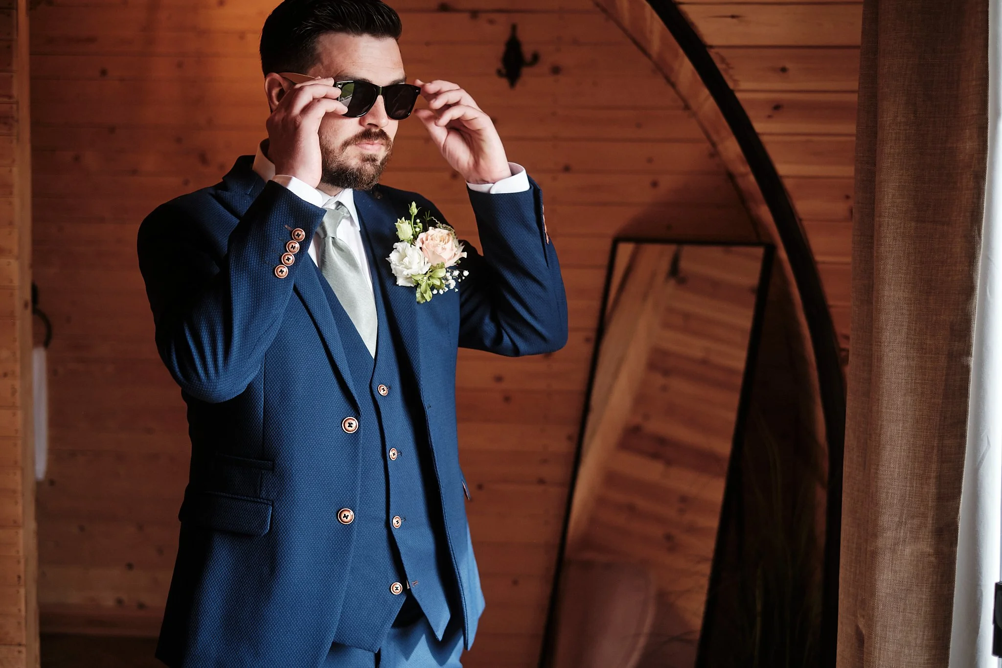 A man wearing a dark blue suit and light gray tie adjusting black sunglasses indoors with wooden walls and a curtain in the background.