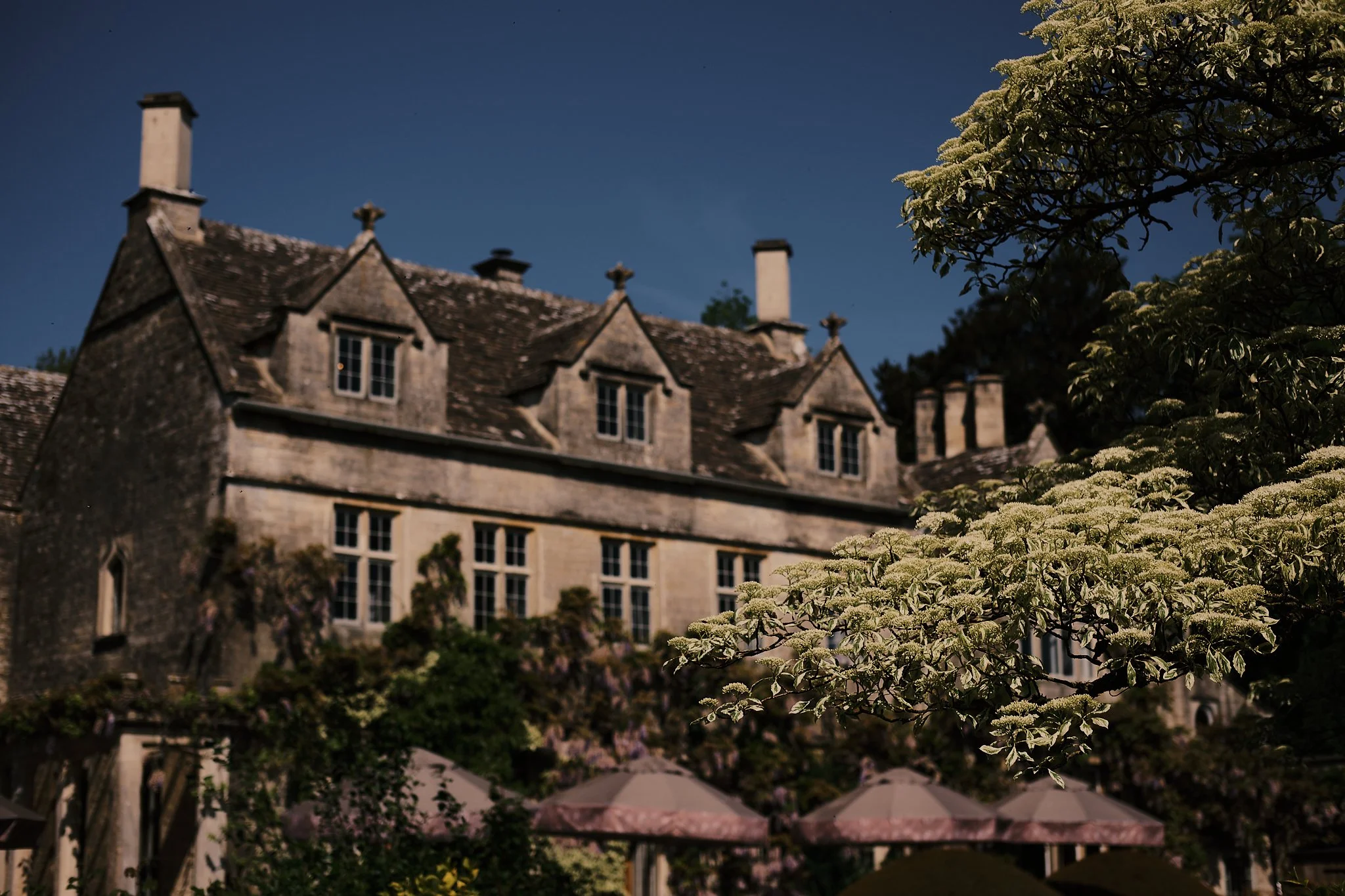 A large historic stone building with multiple chimneys, surrounded by trees and flowering bushes, under a clear dark blue sky.