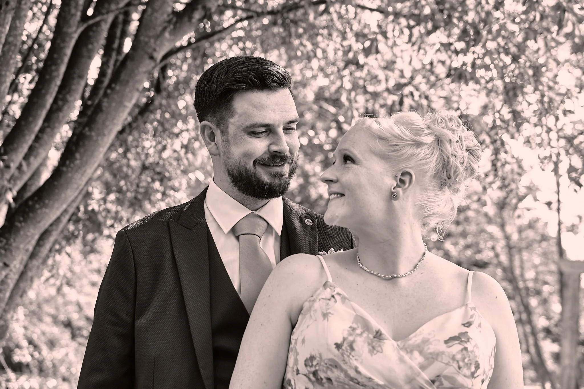 A black and white photo of a man and woman looking at each other outdoors, with trees in the background. The man has dark hair and a beard, wearing a suit and tie. The woman has light hair styled in an updo, wearing a dress with a floral pattern and 