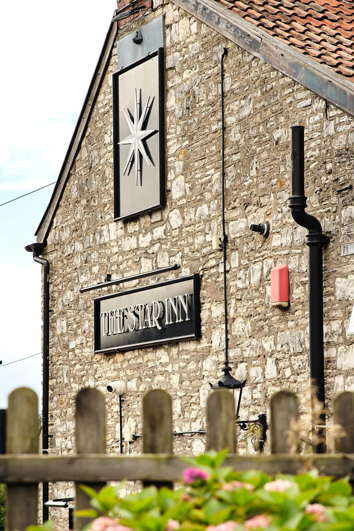 Close-up of the exterior wall of a building with a stone facade, displaying a sign that reads 'The Star Inn' and a clock with a star-shaped design. There are square security cameras, a streetlamp, and black pipes attached to the wall. A wooden fence 