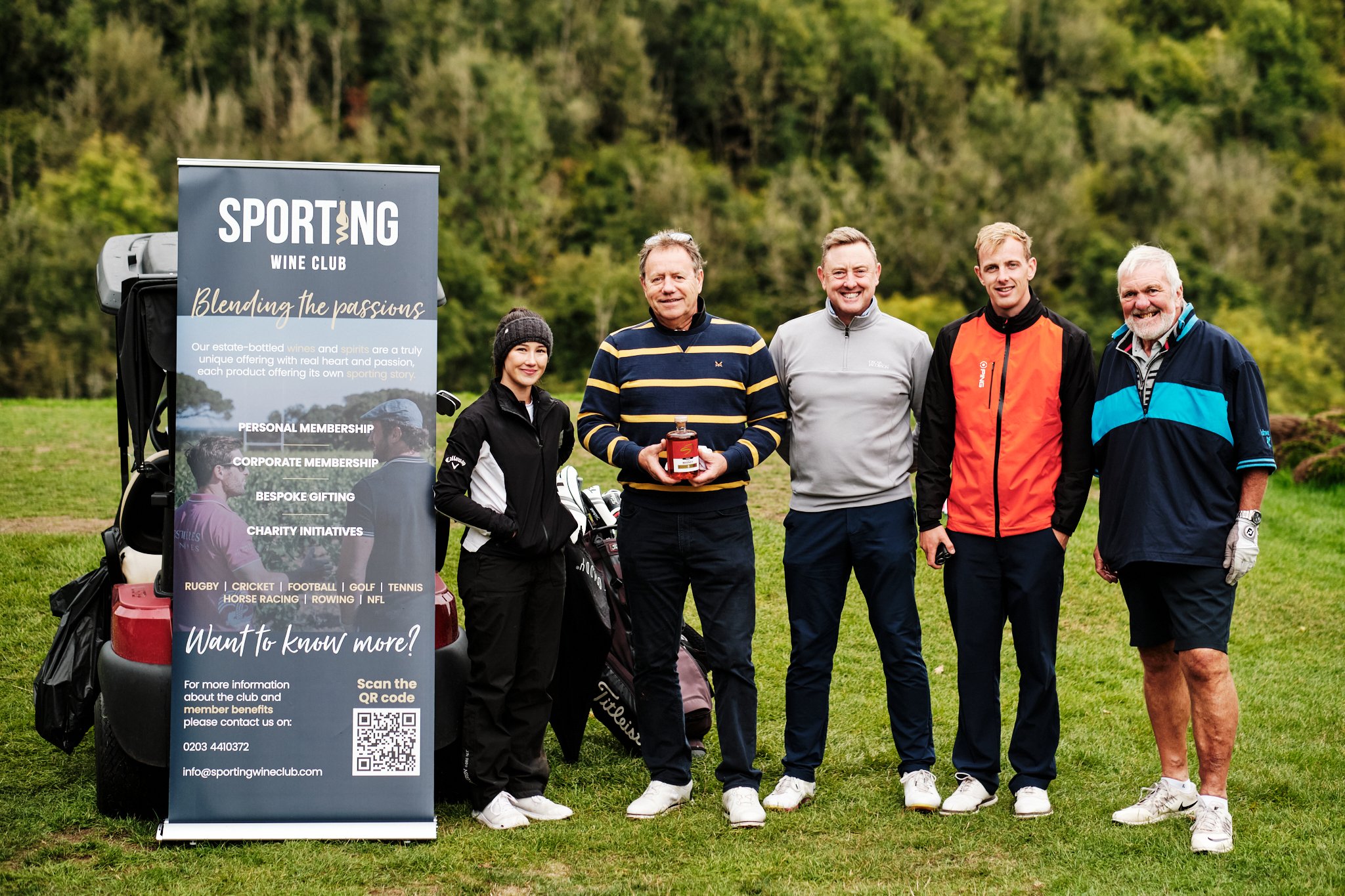 Group of five people standing outdoors at a golf course, holding a bottle of alcohol, next to a banner for Sporting Wine Club, with trees in the background.