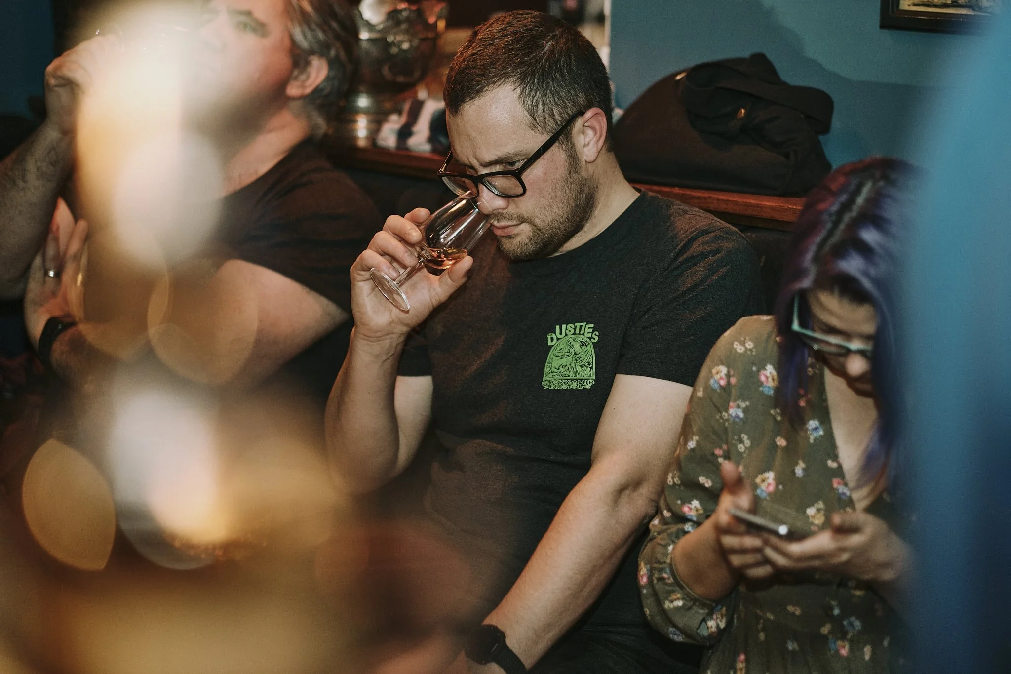 A man with glasses and a dark T-shirt tasting a glass of rosé wine while sitting next to a woman with purple hair and glasses looking at her phone, in a dimly lit setting.