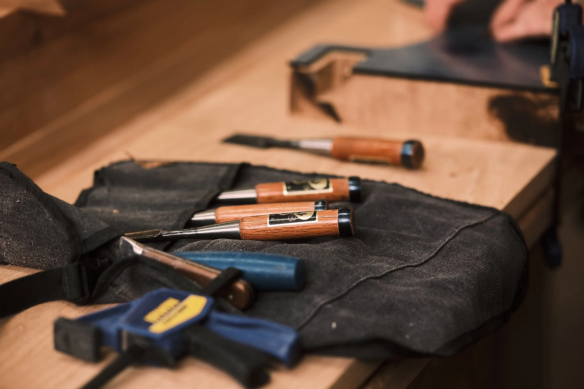 Wood carving chisels with wooden handles in a black tool roll on a wooden workbench.
