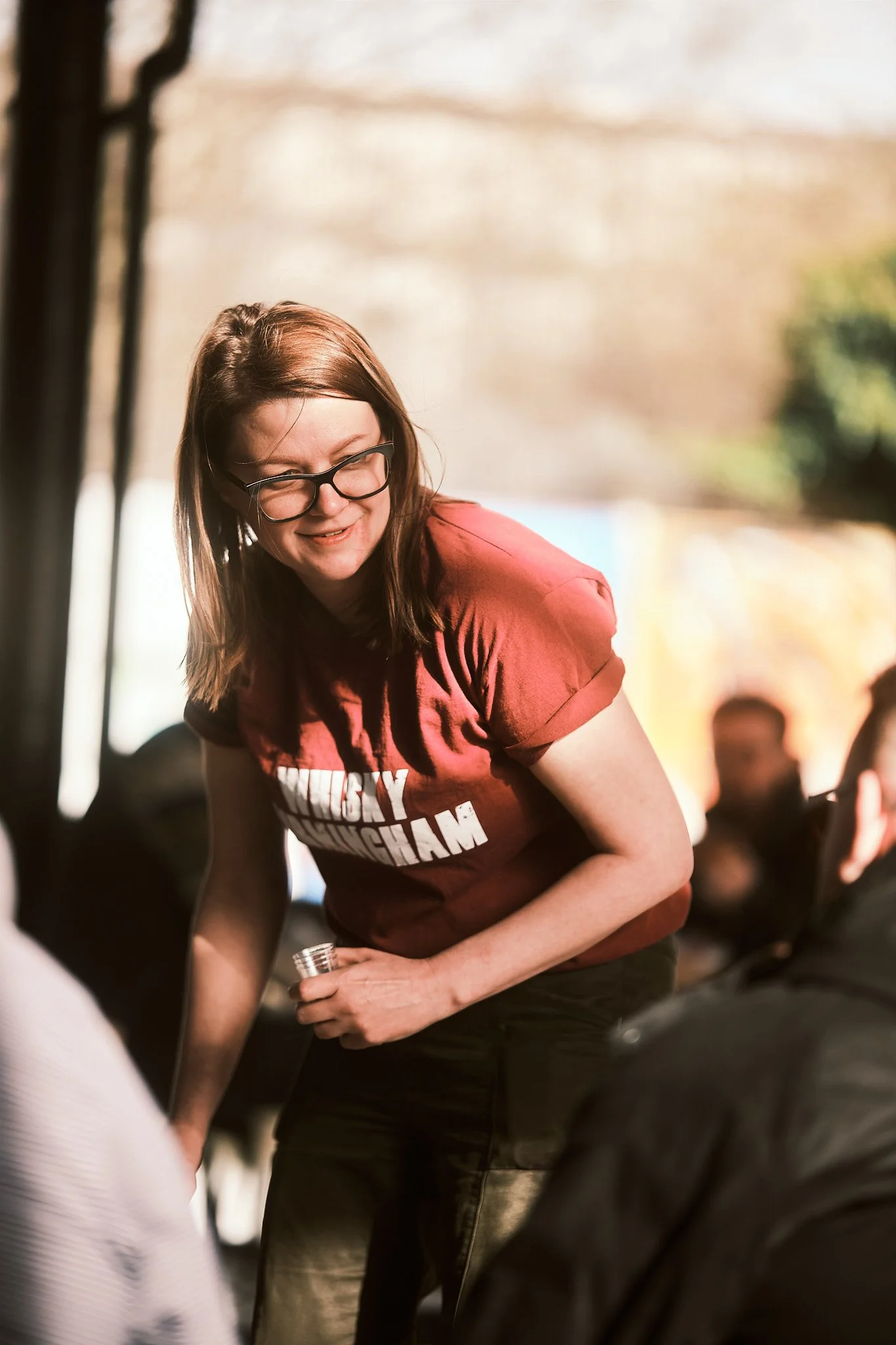 A woman wearing glasses and a red T-shirt with text is smiling and engaged in conversation at an indoor event.