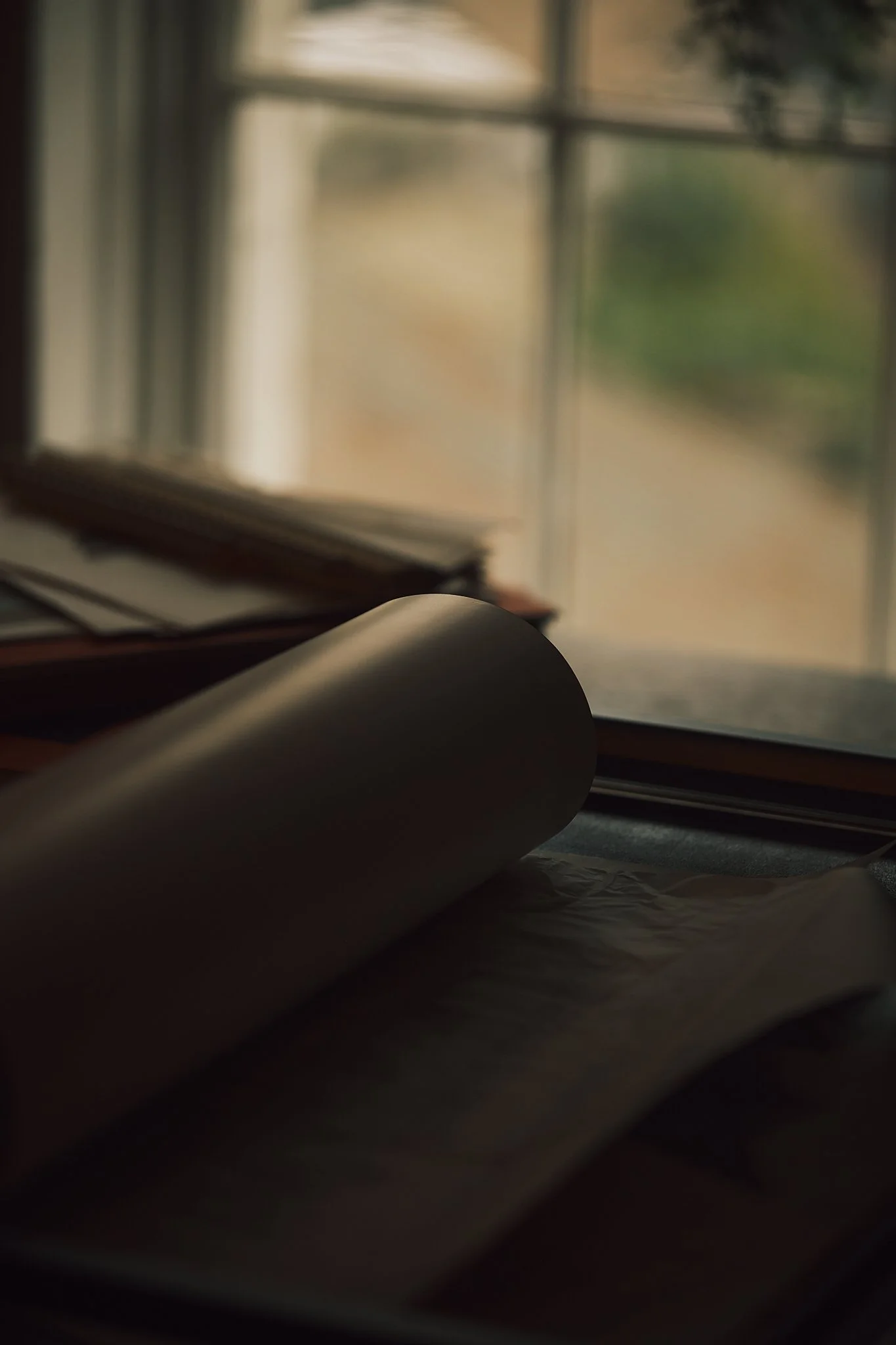 Close-up of a black rolled-up paper or poster on a table near a window, with blurred papers and a window showing a soft outdoor view in the background.