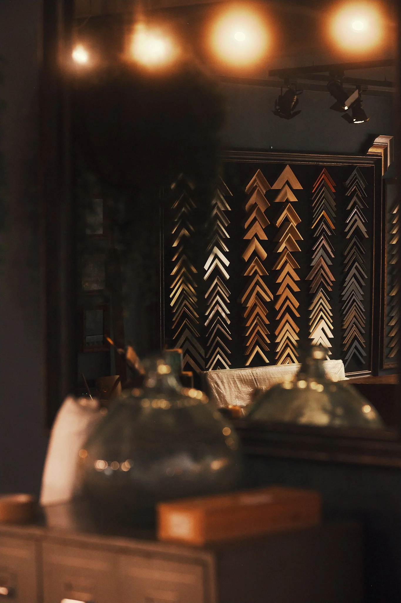A cozy, warmly lit room with a dark wall decorated with a pattern of small wooden triangles arranged in vertical rows, illuminated by soft overhead lights. In the foreground, there are blurred glass cloches and a wooden object on a table or counter.