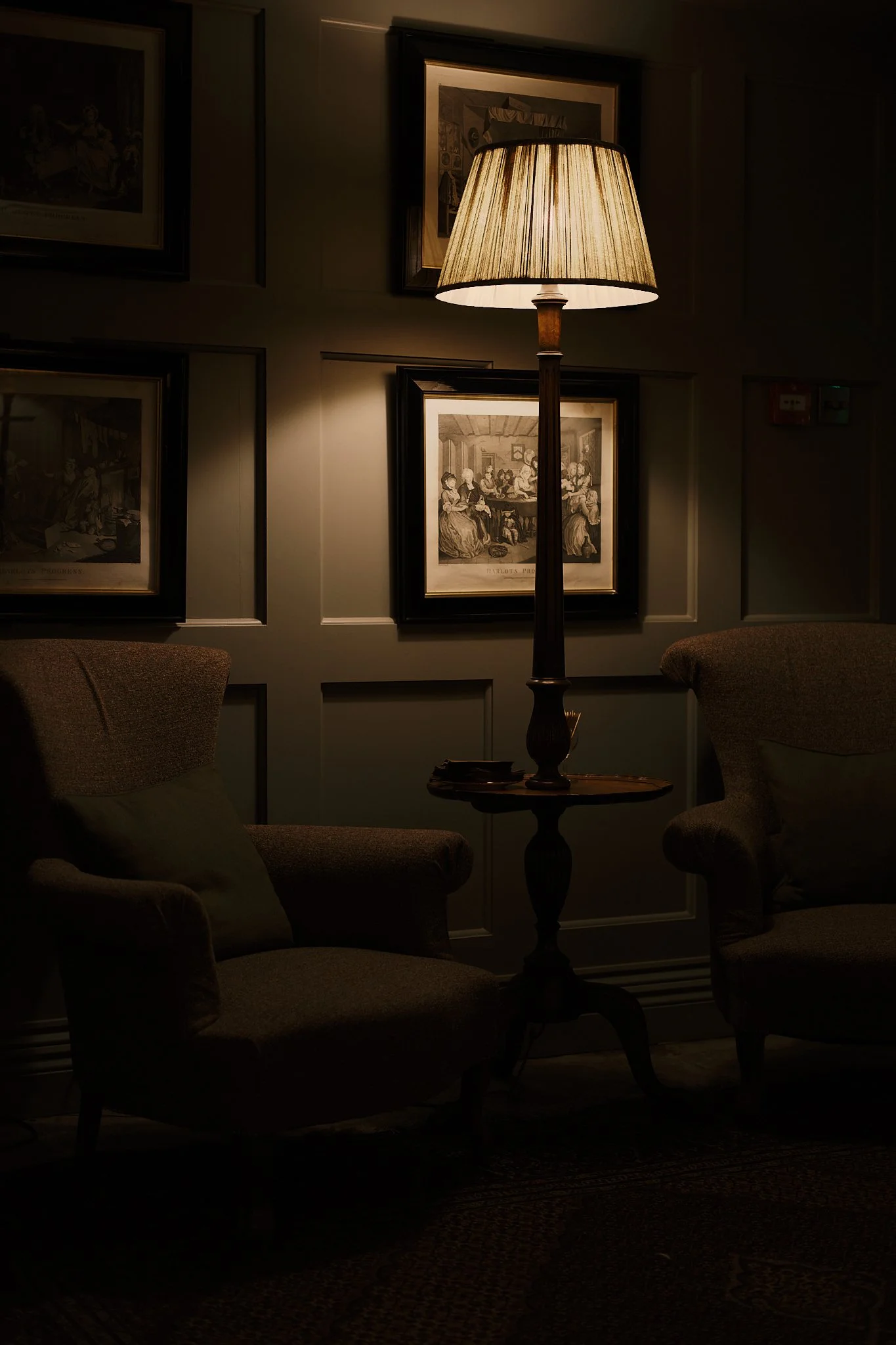 A dimly lit room with two beige armchairs, a wooden side table, framed artwork on the wall, and a tall floor lamp with a beige lampshade casting warm light. Hotel interior photography at THE PIG-in the Cotswolds