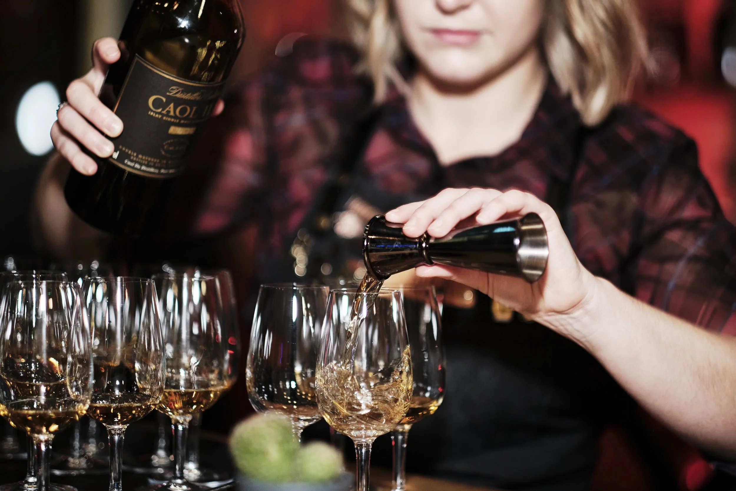 A woman pouring whiskey into a glass at a bar, with more glasses and a bottle of whiskey on the counter.