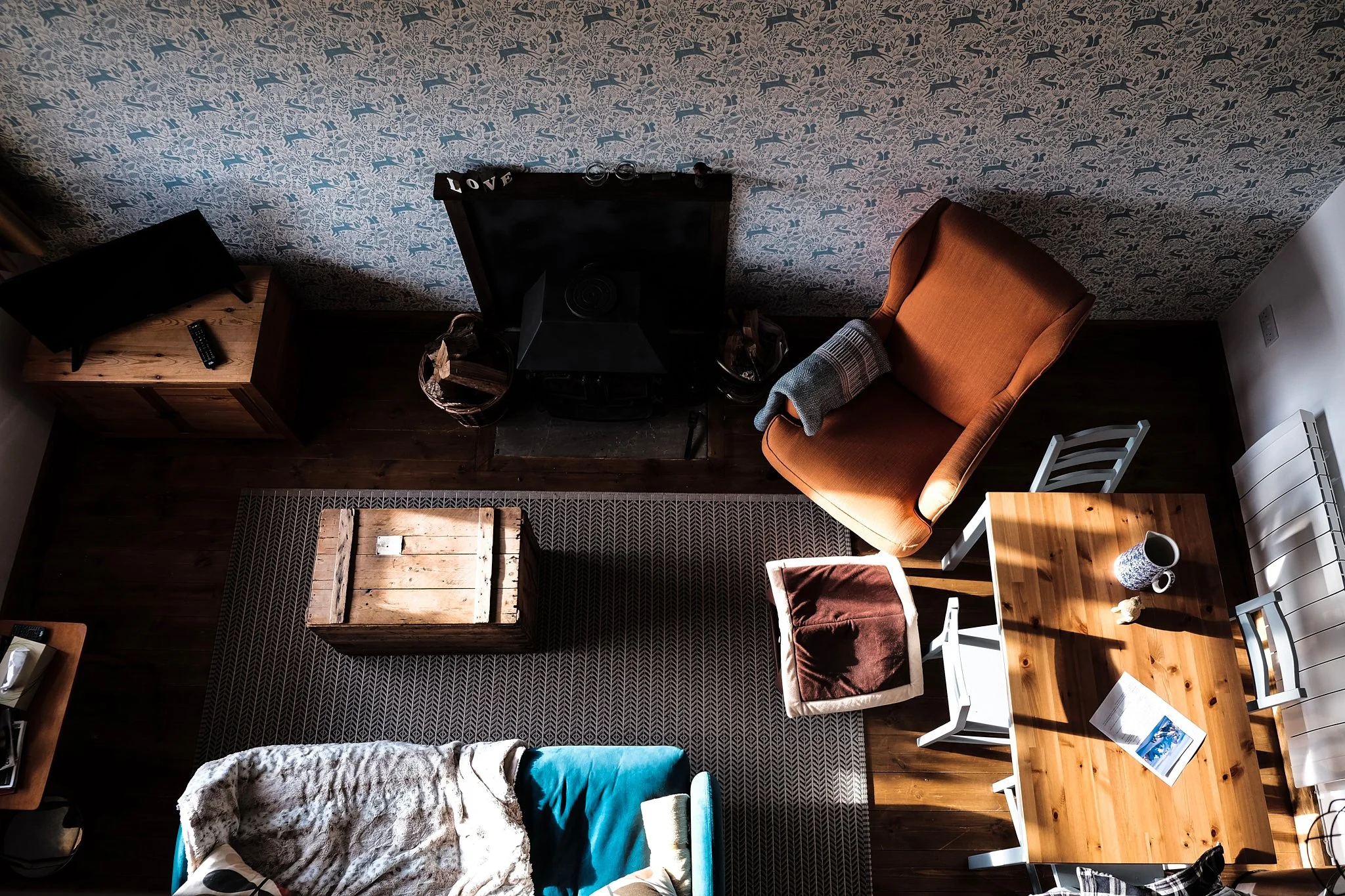 Top-down view of a cozy living room with a patterned blue wallpaper, wooden furniture, an orange armchair with a blanket, a wooden coffee table. A sofa with a blanket, a chair, a small TV on a wooden stand, and a dining table with chairs are visible.