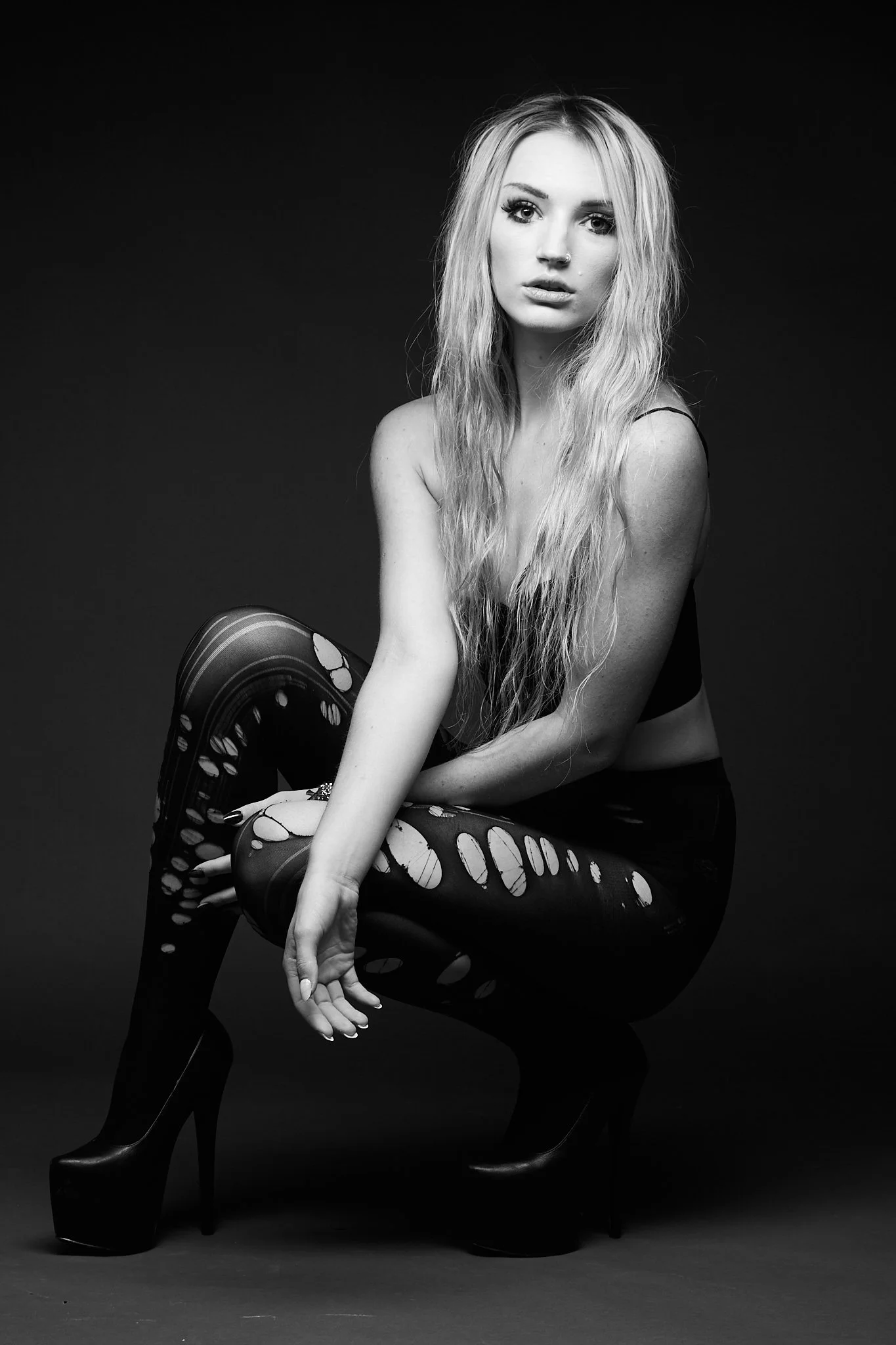 Black and white studio portrait of a young woman with long, wavy blonde hair, sitting down with one knee raised and wearing patterned leggings and high heels.