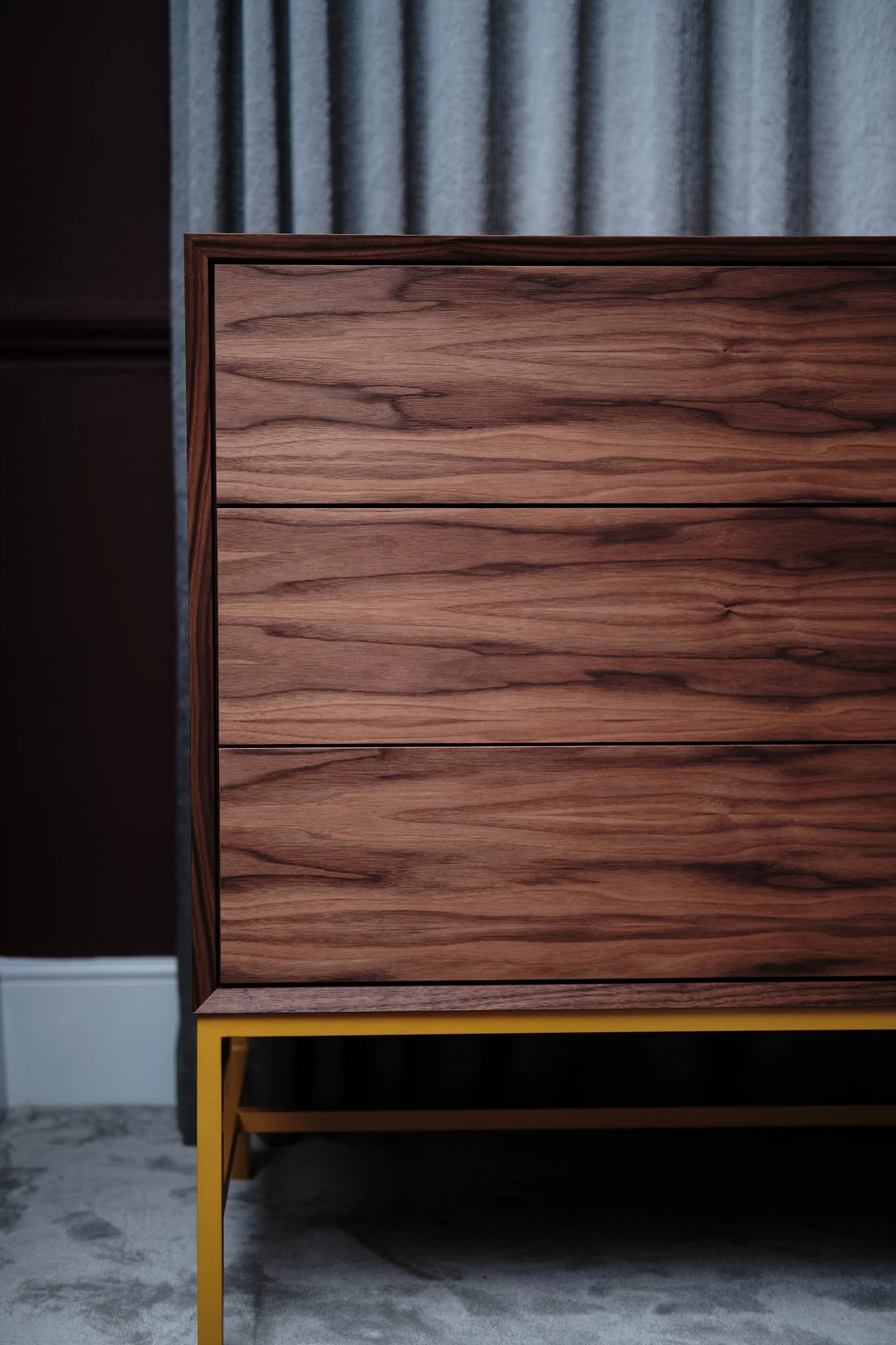 Close-up of a wooden dresser with three drawers, set against a dark wall and a textured gray curtain in the background, with a yellow metal frame supporting the dresser.