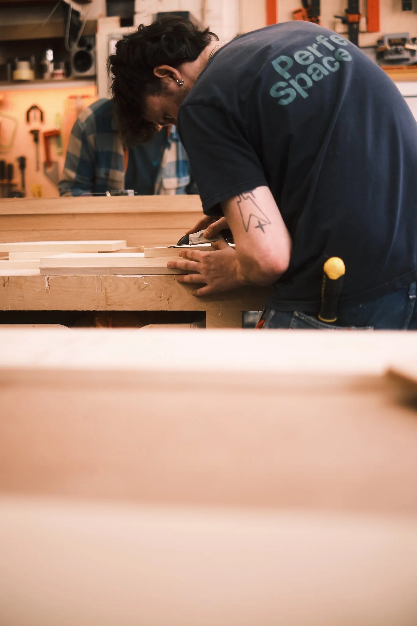 A person woodworking in a shop, focused on sawing a piece of wood, with tools and workbenches visible in the background.