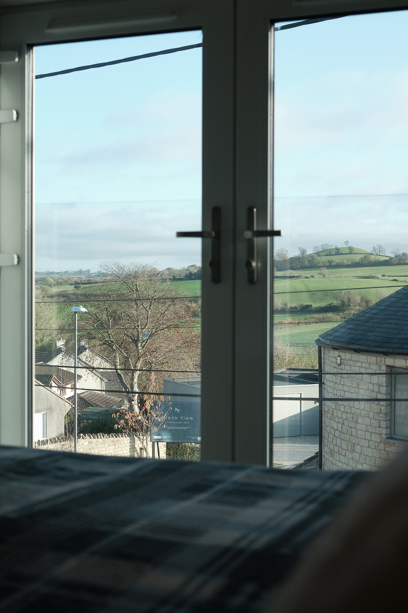 View through a window showing a rural landscape with green hills, a tree, and some houses and buildings.