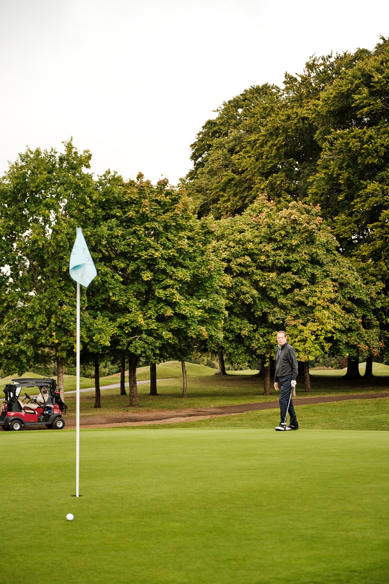 A man preparing to putt on a golf course, with a golf cart in the background and trees surrounding the green.