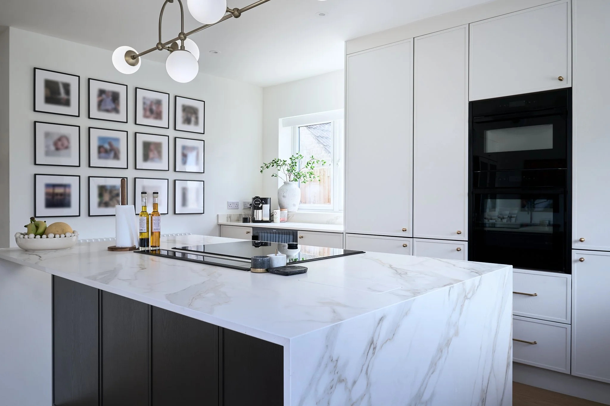 Modern white kitchen with marble island, black built-in oven, wall of cabinets, and a window with a plant on the counter.