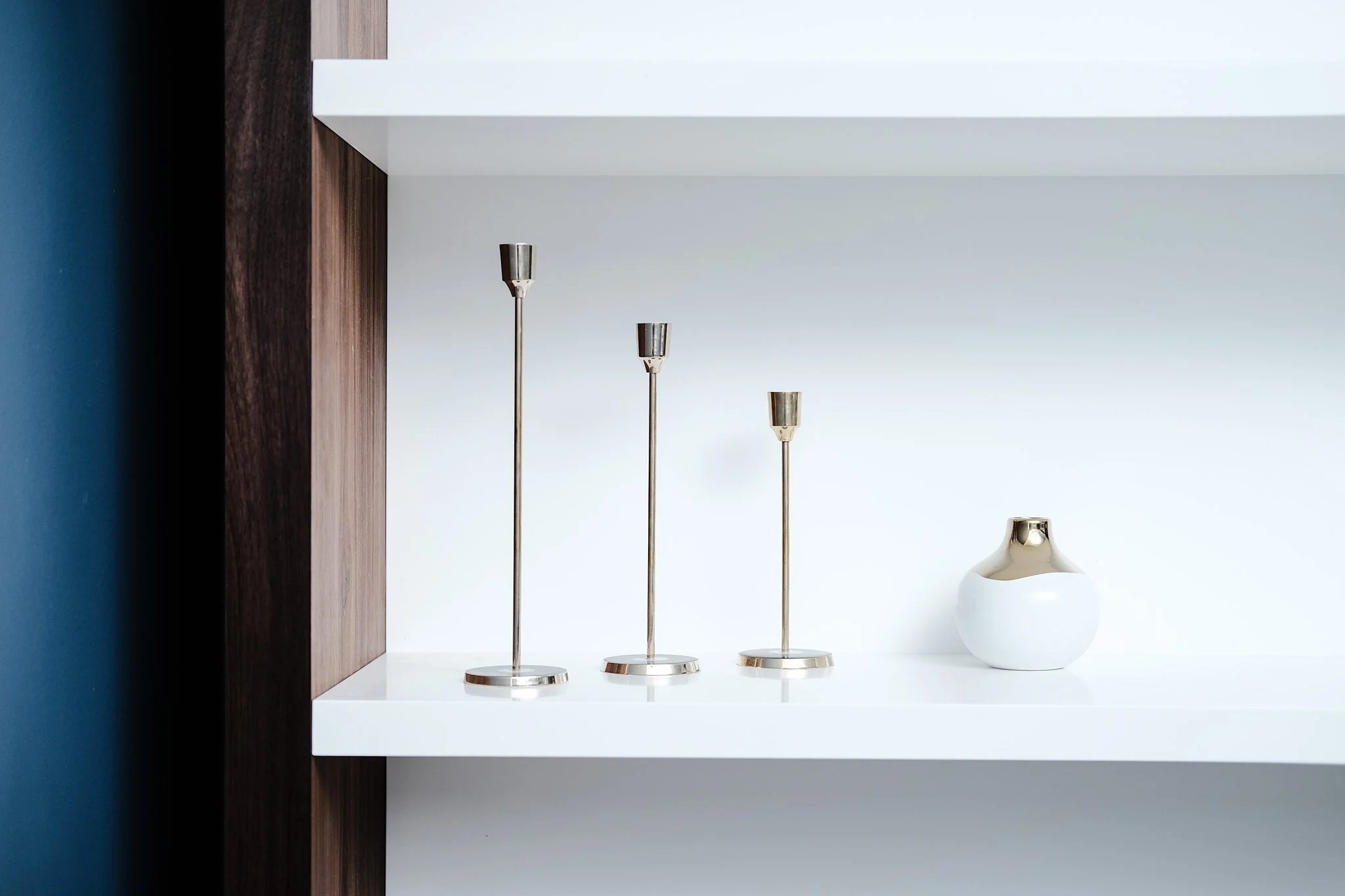 Three modern silver candle holders of varying heights and a white and silver decorative vase on a white shelf with a dark wooden side and a blue wall in the background.