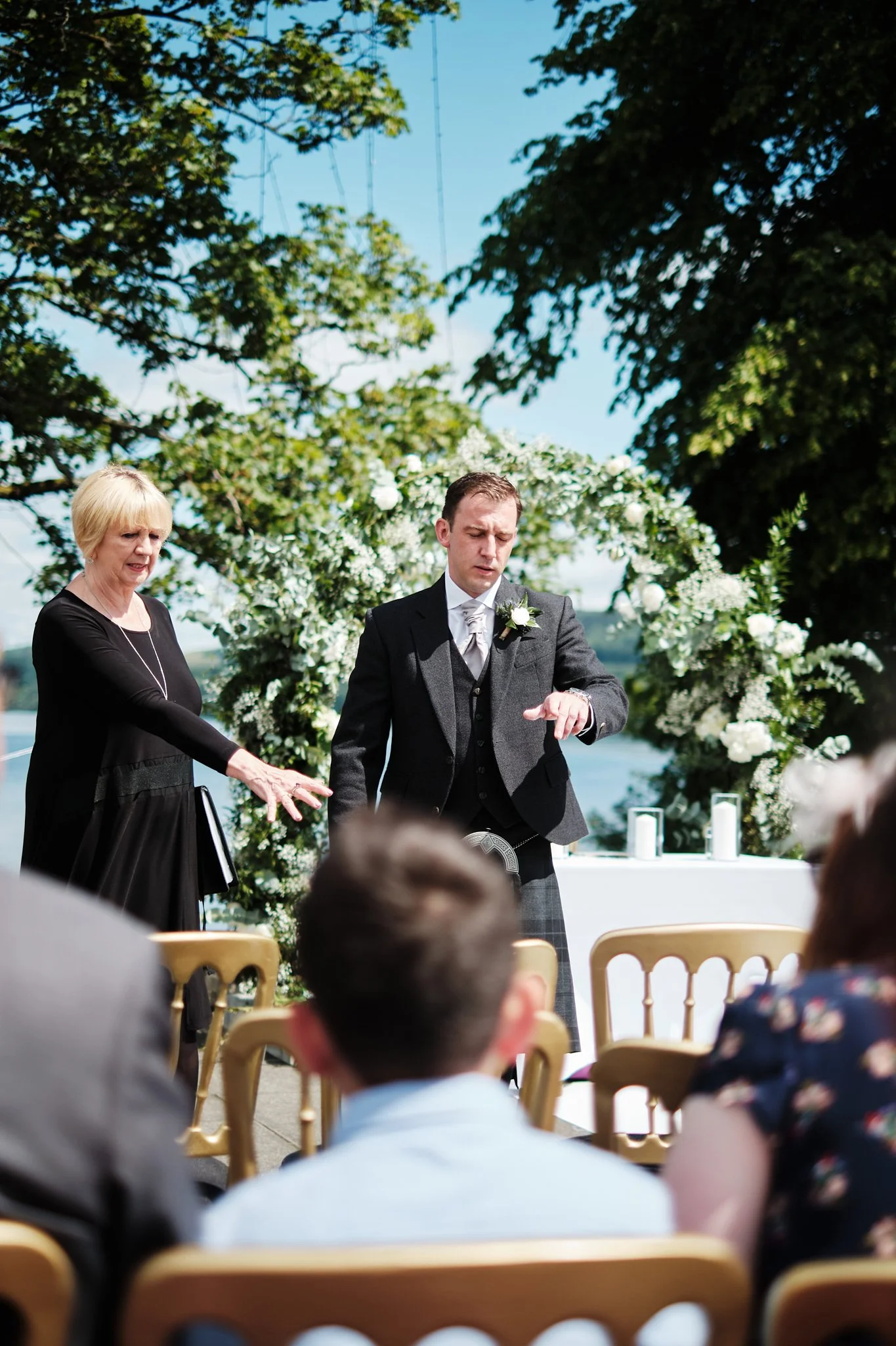 A wedding ceremony outdoors on a sunny day, with a bride and groom standing in front of an arch decorated with white flowers, with guests seated on chairs watching the ceremony.